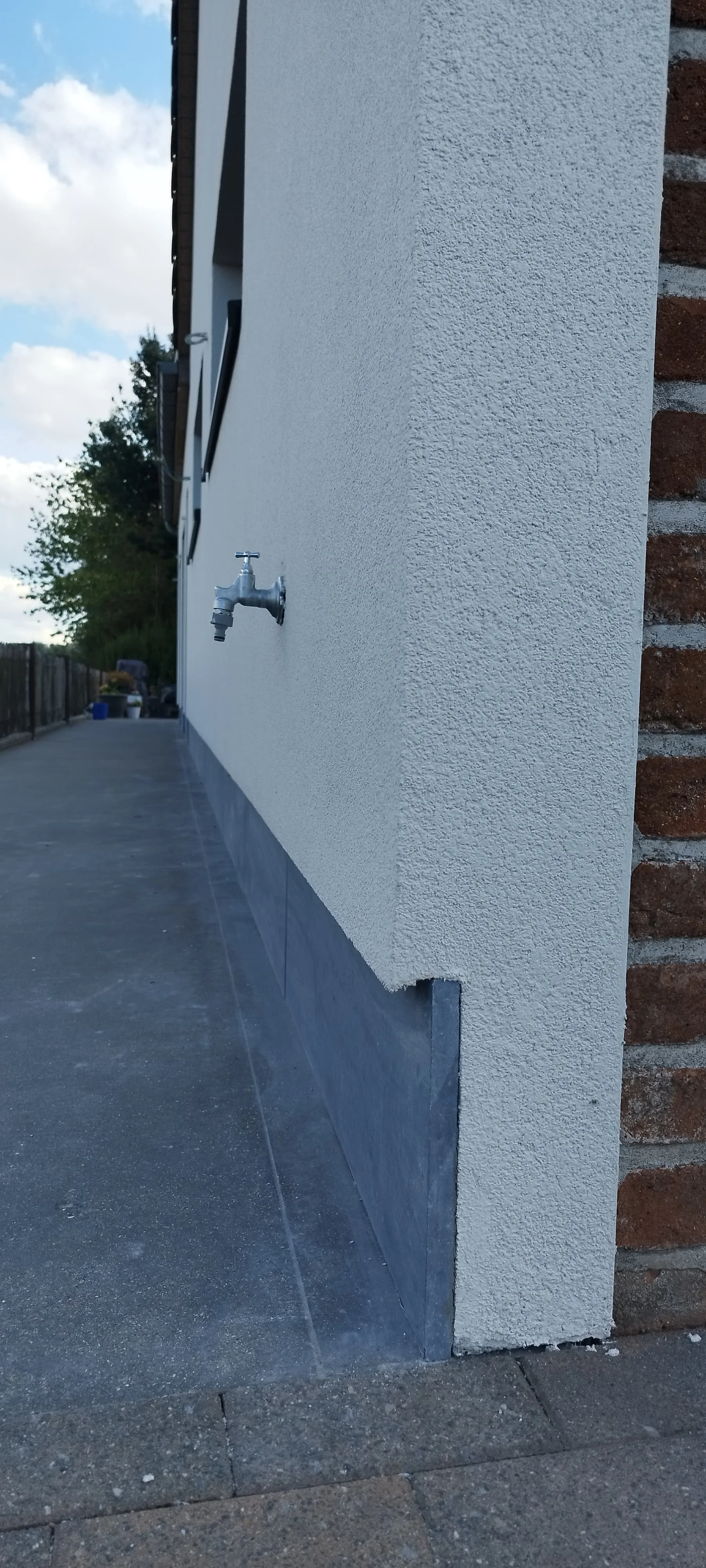 Buitenzijde van een huis met een waterkraan en een begroeide tuin op de achtergrond.