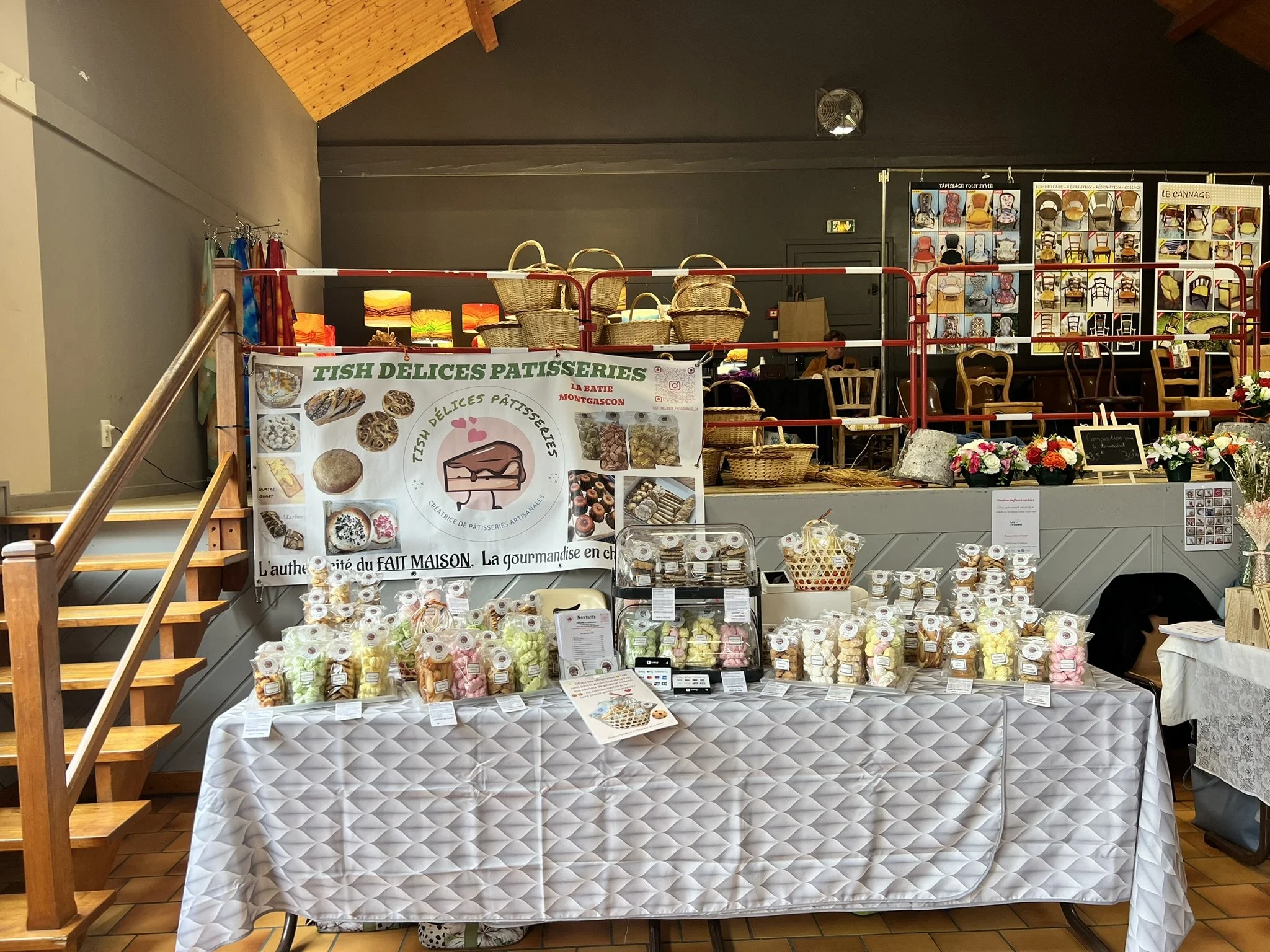 Un stand de vente de pâtisseries artisanales, avec un panneau indiquant 'TISH DELICES PATISSERIES', présentant divers bonbons, pâtisseries, et biscuits emballés, avec des fleurs et chaises en arrière-plan.