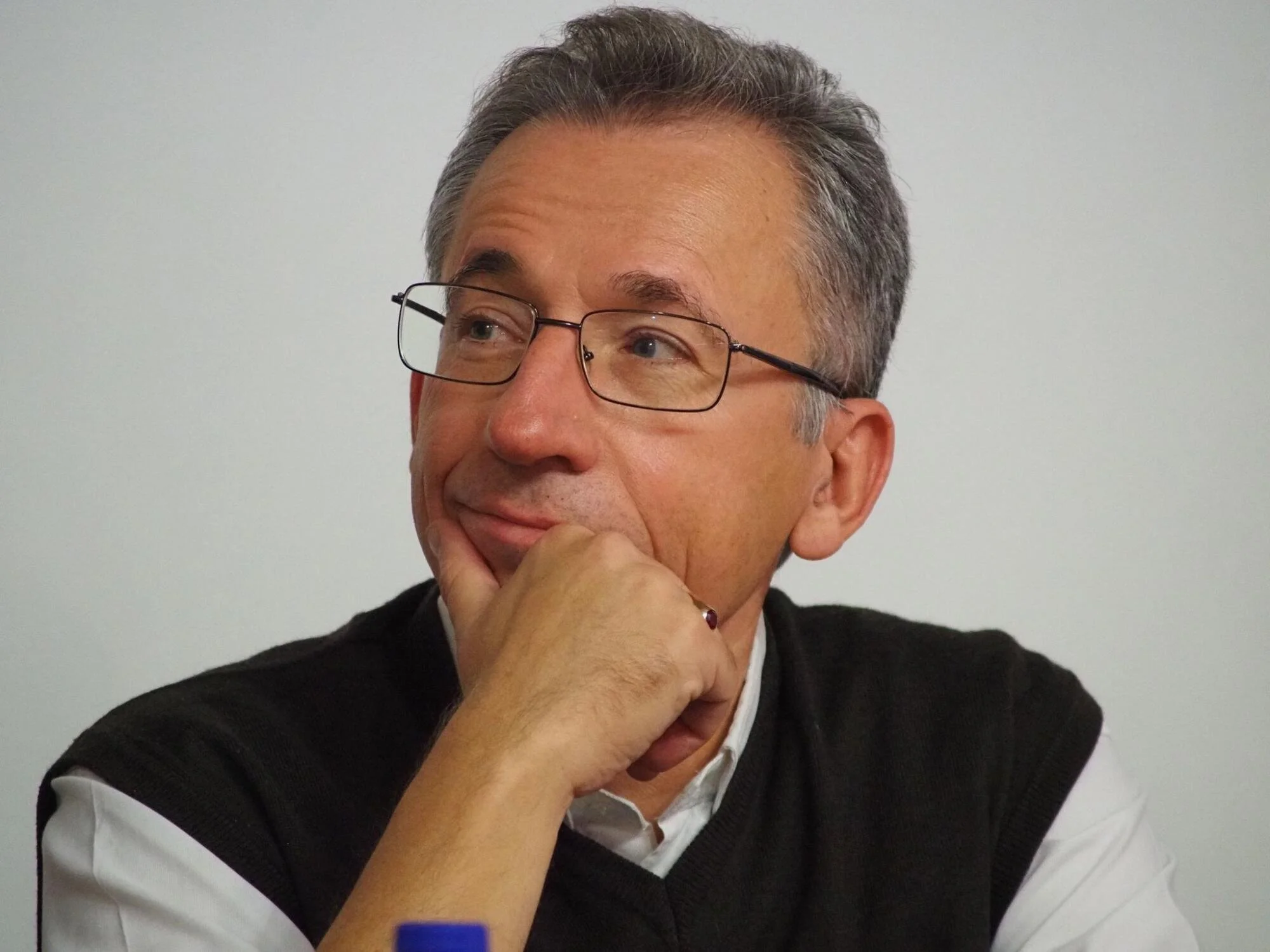 A middle-aged man with gray hair and glasses, wearing a white shirt and black sweater, appears to be in thought, with his hand resting on his chin and looking to the side.
