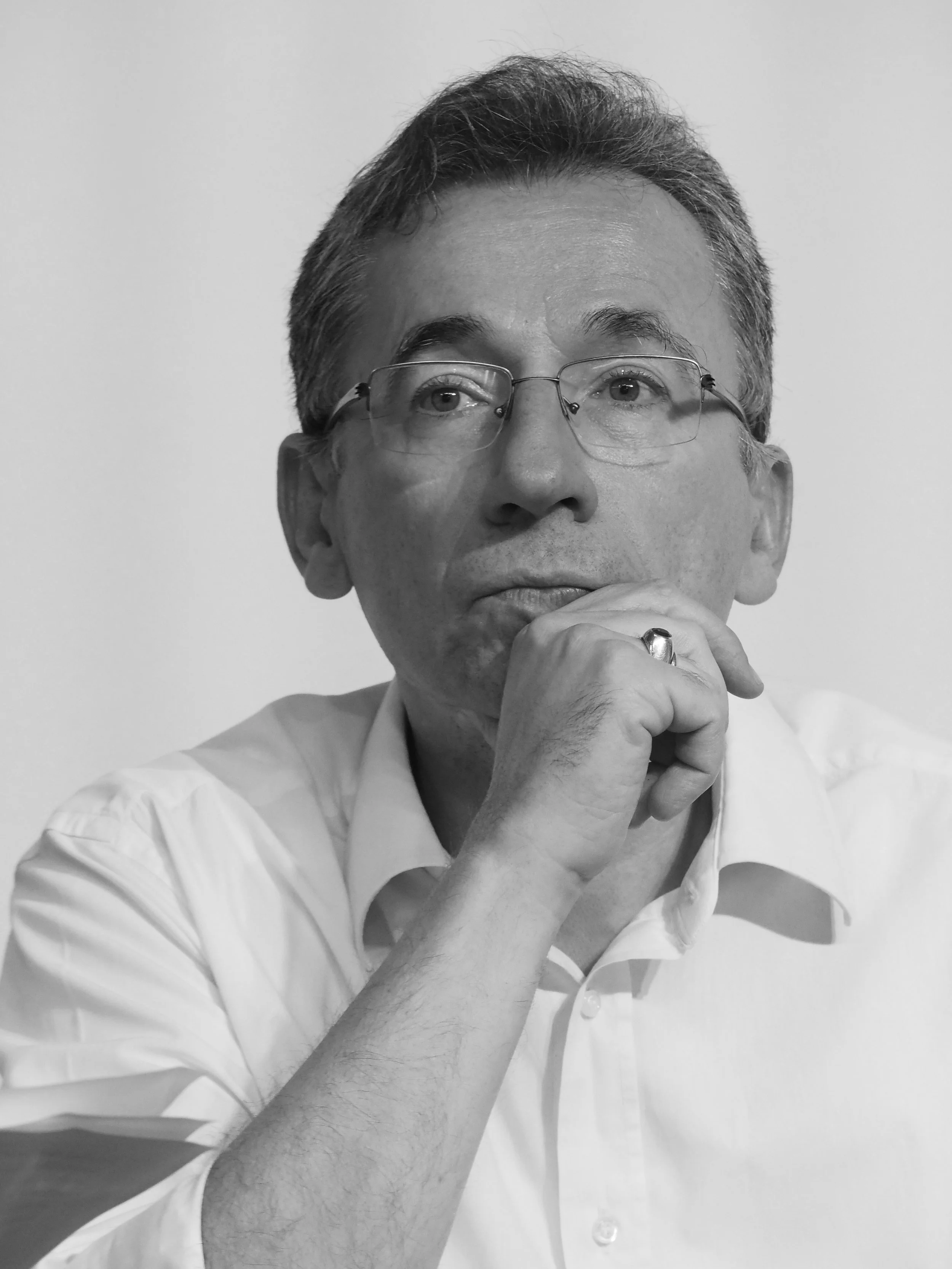 A black and white portrait of Isidoros Zourgos wearing glasses, with short, slightly disheveled hair, holding his chin with his right hand, and looking slightly upward.