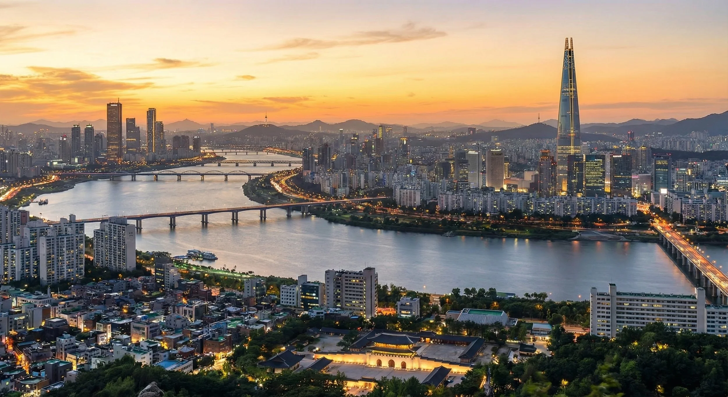 A city skyline at sunset with a river running through the city, numerous bridges crossing the river, modern high-rise buildings, and mountains in the background.
