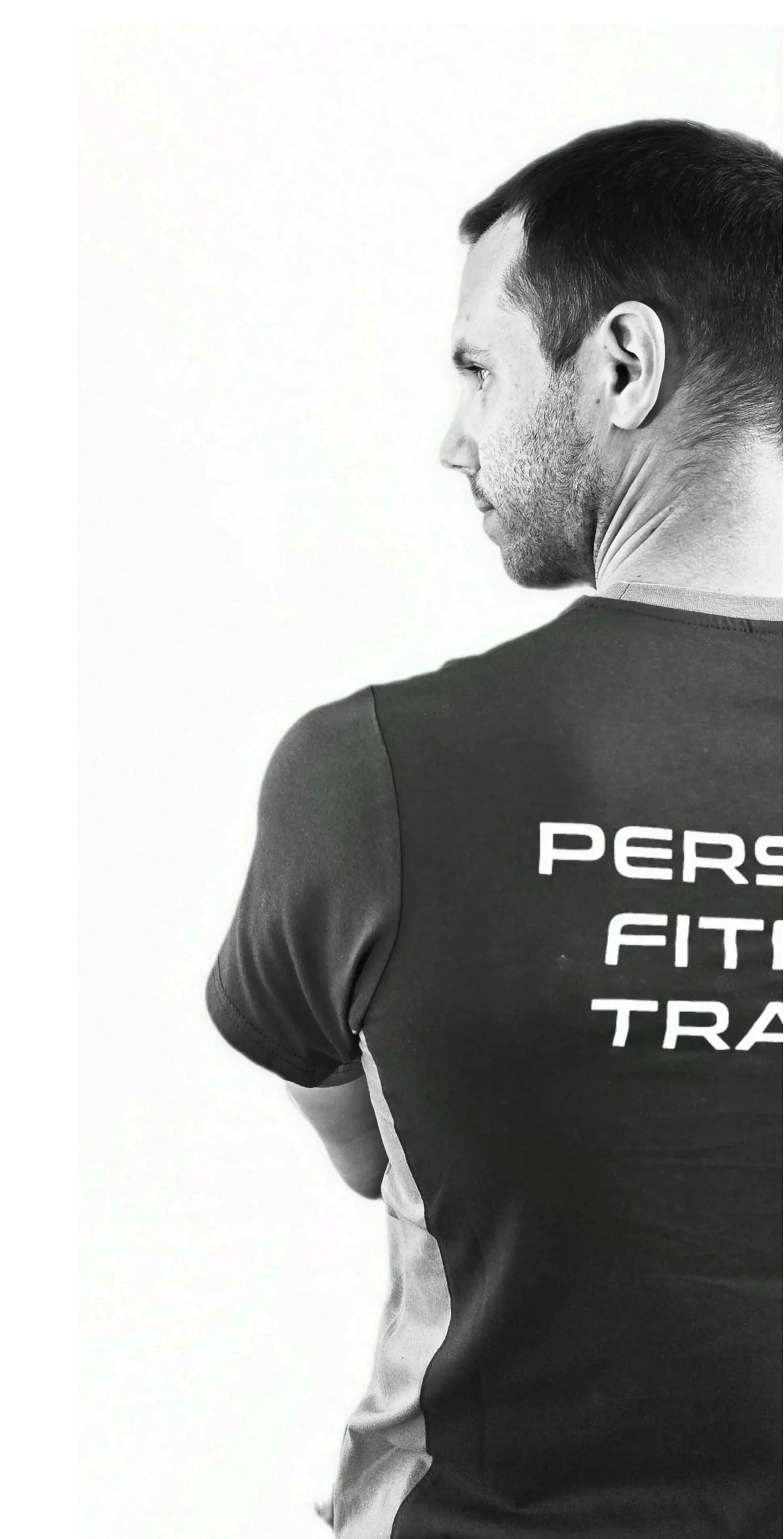 Un uomo in maglietta sportiva nera con scritto 'Personal Fitness Training' vista di schiena, con capelli scuri e barba leggera, posa di profilo contro uno sfondo bianco.