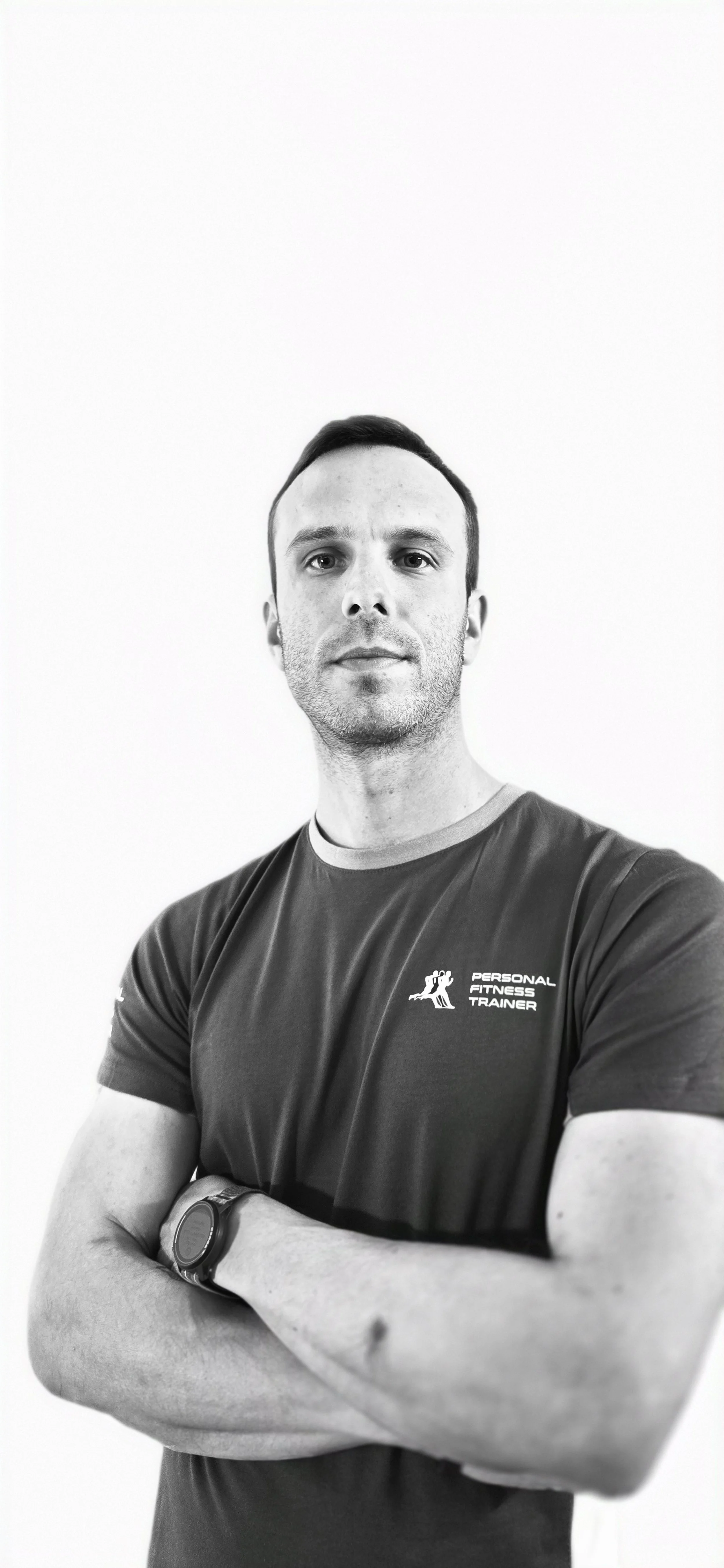 Uomo con braccia incrociate indossa una maglietta con logo fitness e ha un orologio al polso, posa con sfondo bianco.