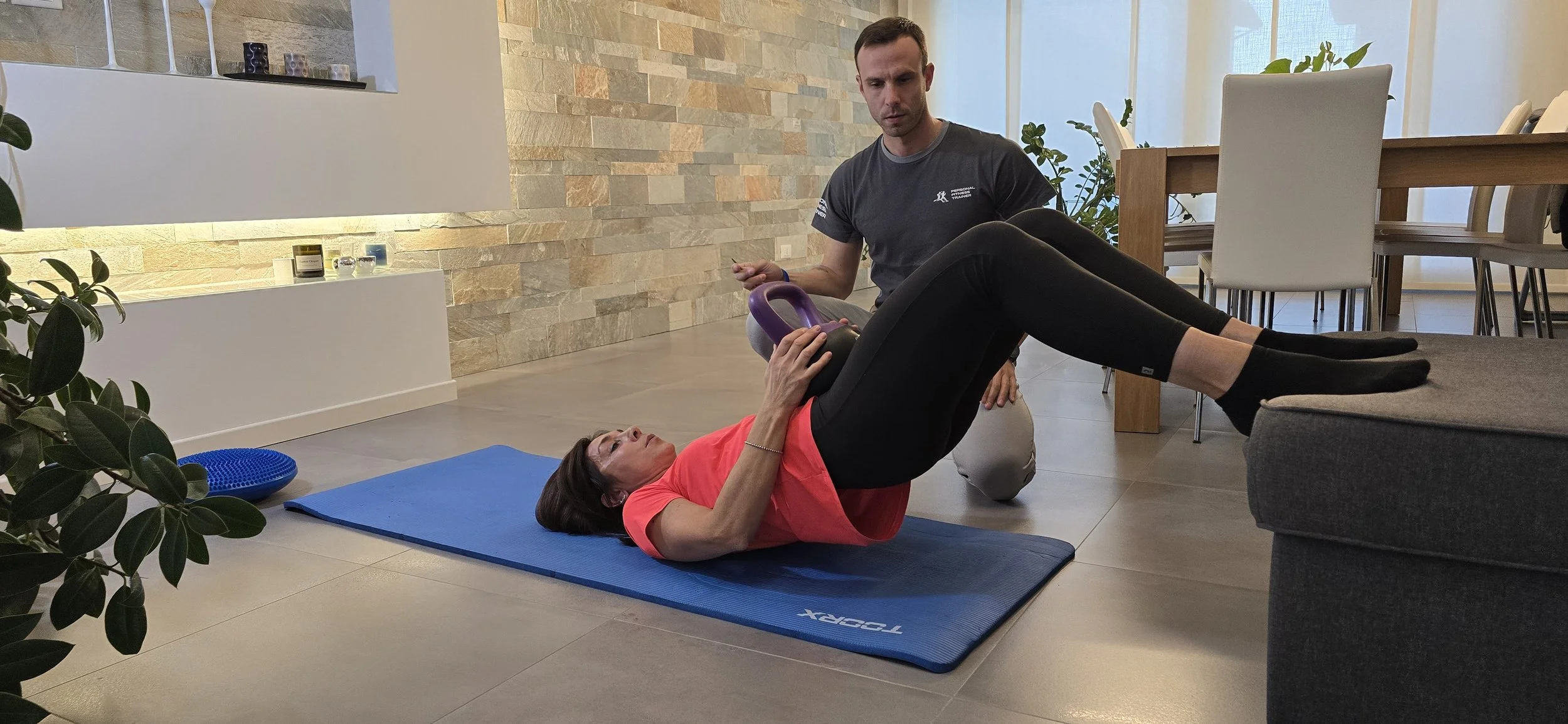 Una donna fa esercizi con un kettlebell su un tappetino blu, assistita da un istruttore in un ambiente domestico.