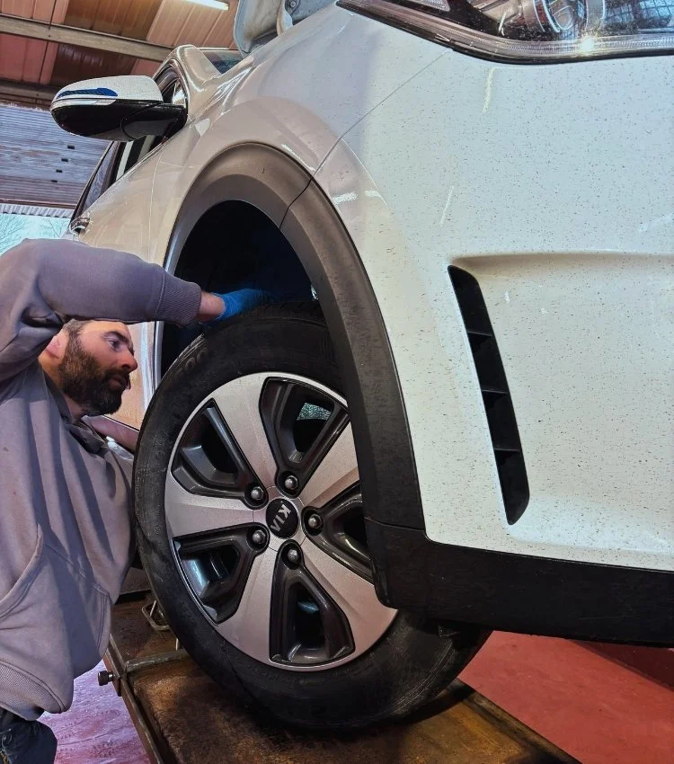 A man wearing a gray hoodie and blue gloves inspecting the wheel of a white Kia vehicle in a garage.