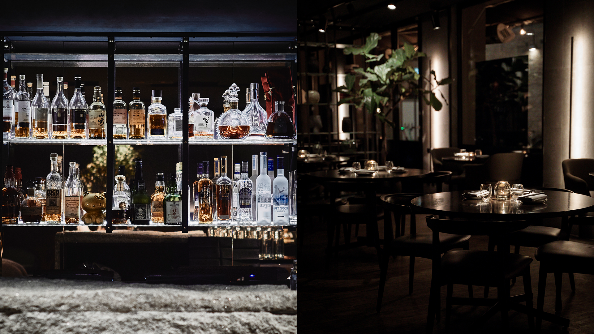 A dimly lit restaurant interior with round tables, glasses, and tableware, with a bar on the left side displaying various bottles of alcohol.