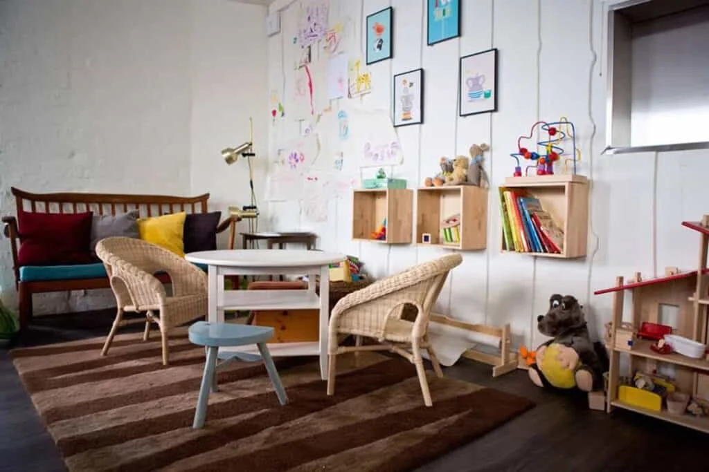 A cozy playroom with a wooden bench with colorful pillows, a small table with chairs, wall-mounted shelves with books and toys, and children's drawings on the wall. There are stuffed animals and a toy garage on the floor.