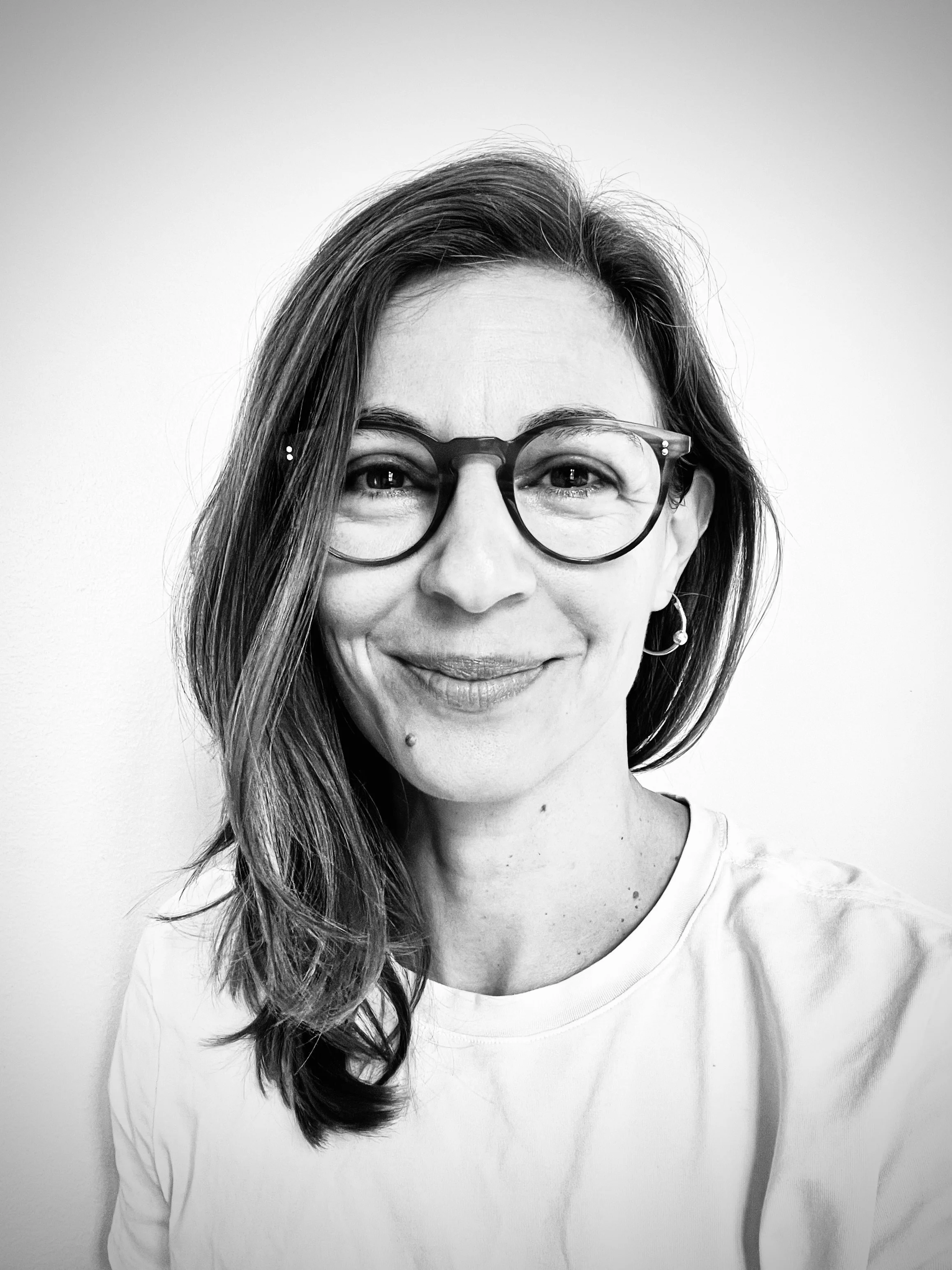 Black and white portrait of a woman with long hair, glasses, and earrings, smiling at the camera against a plain background.