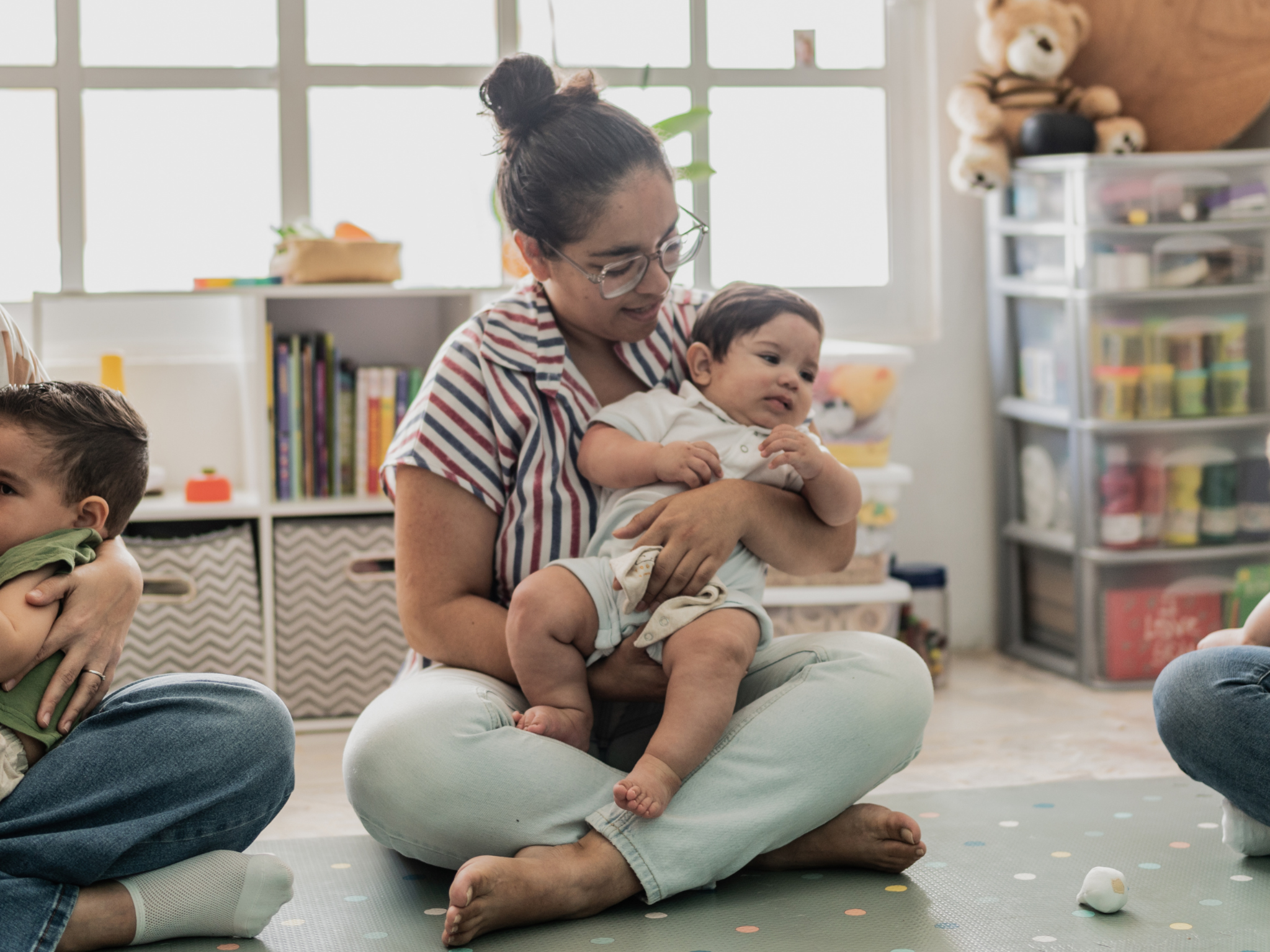 A woman with glasses sitting on the floor, holding a toddler in her lap. The children are engaged, with a bookshelf, storage bins, and a teddy bear in the background.