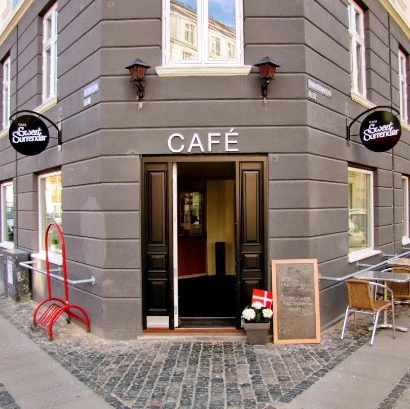 Exterior of a café with black double doors, two black wall-mounted lanterns, and a sign that says 'CAFÉ' above the entrance. There are outdoor tables and chairs, a chalkboard menu, and decorative flowers outside.