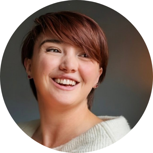 Smiling woman with short brown hair and earrings, wearing a white sweater, against a plain background.