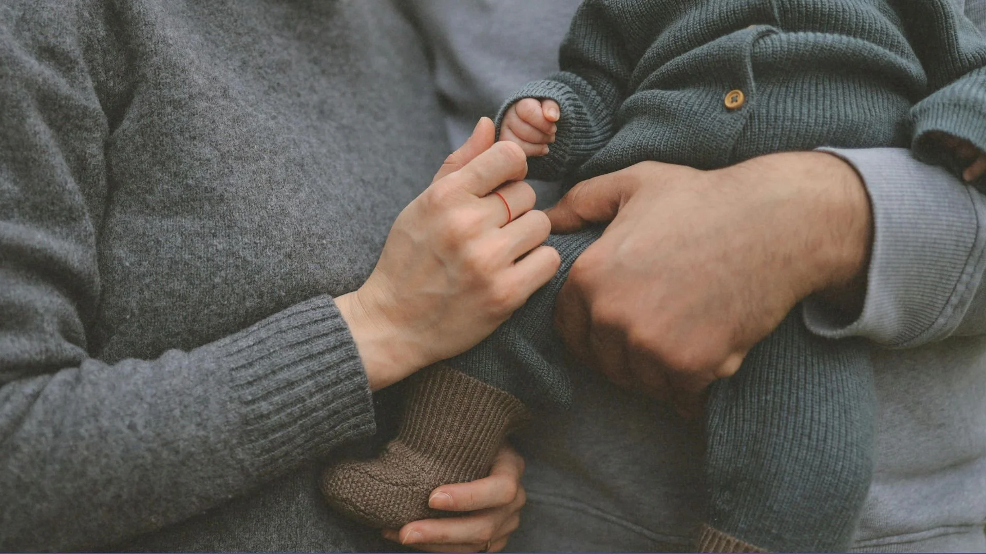 Close-up of two adults holding a baby's hand. The adults are wearing long-sleeved sweaters, one in gray and the other in dark gray, and the baby is dressed in a dark gray knitted sweater and brown pants.