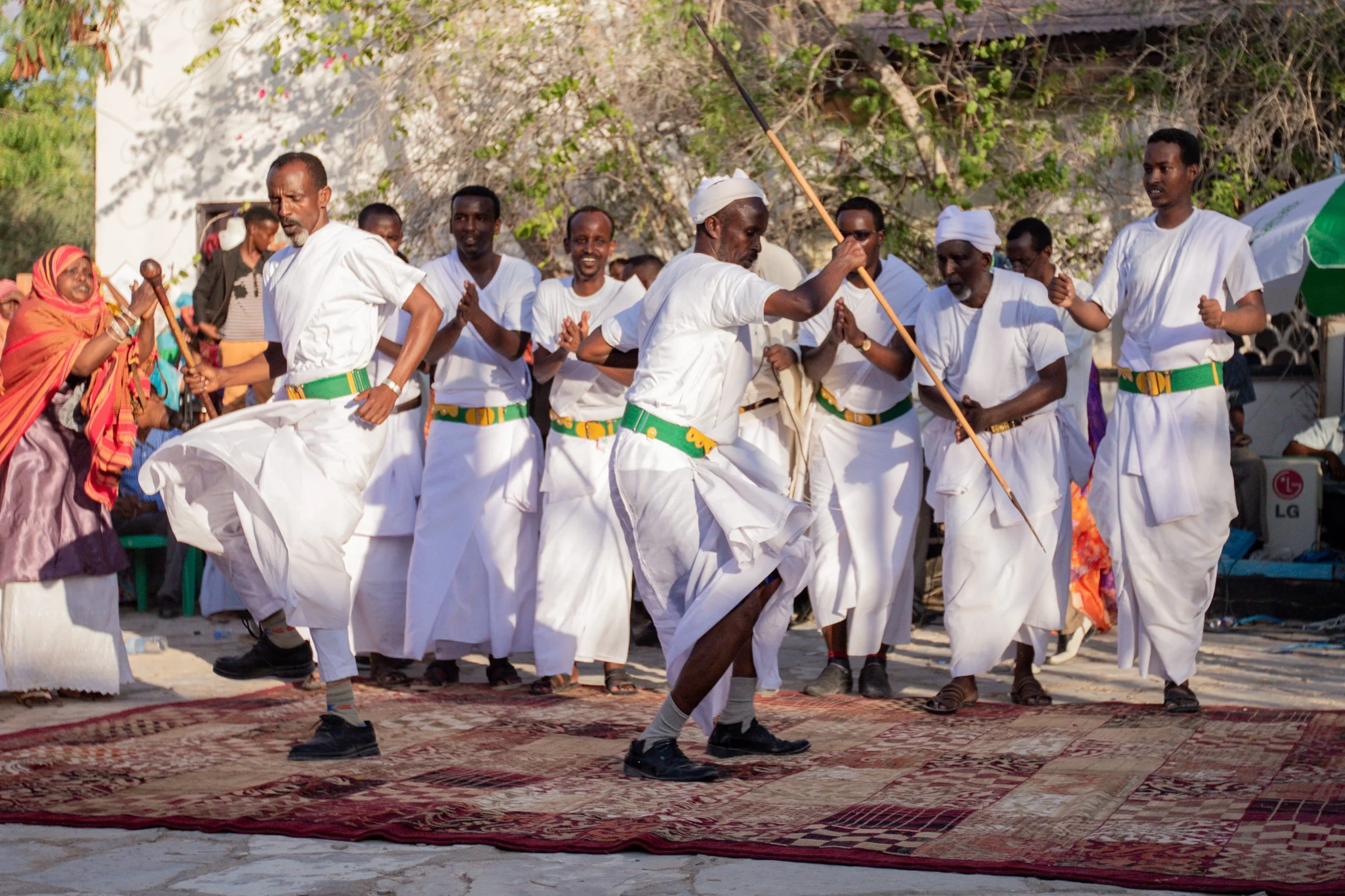 Men dressed in white traditional clothing and green belts dancing on a patterned rug outdoors during a cultural event.