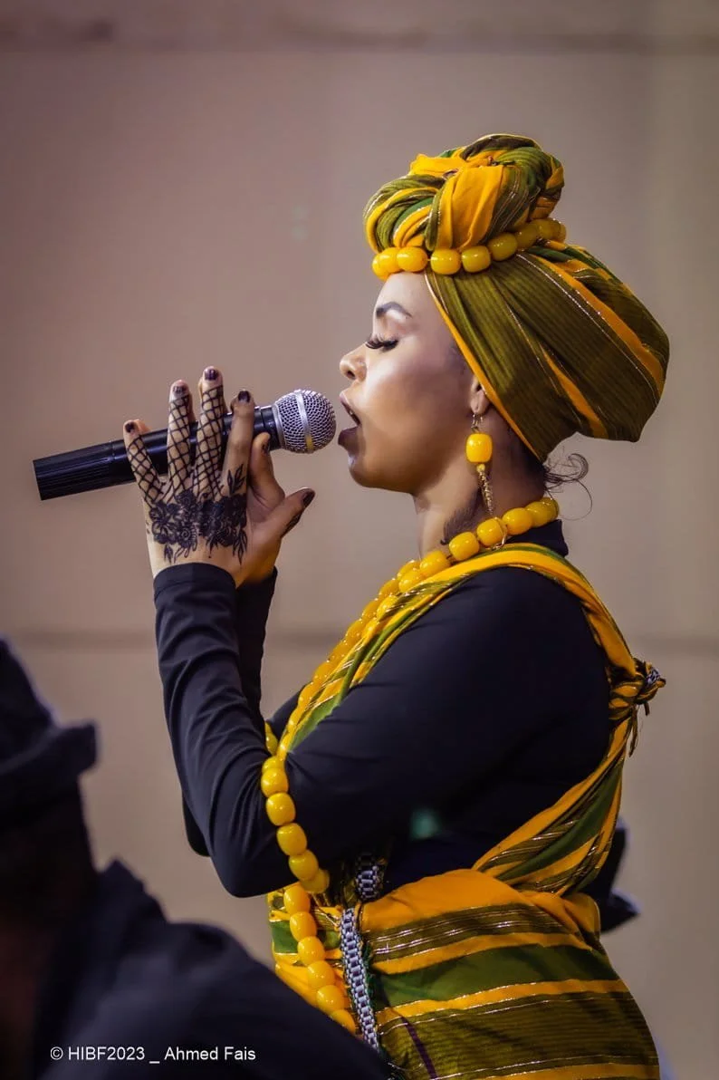 A woman wearing traditional African attire with yellow and green fabric and accessories, singing into a microphone with her eyes closed. She has henna on her hand, a headwrap, and matching jewelry.