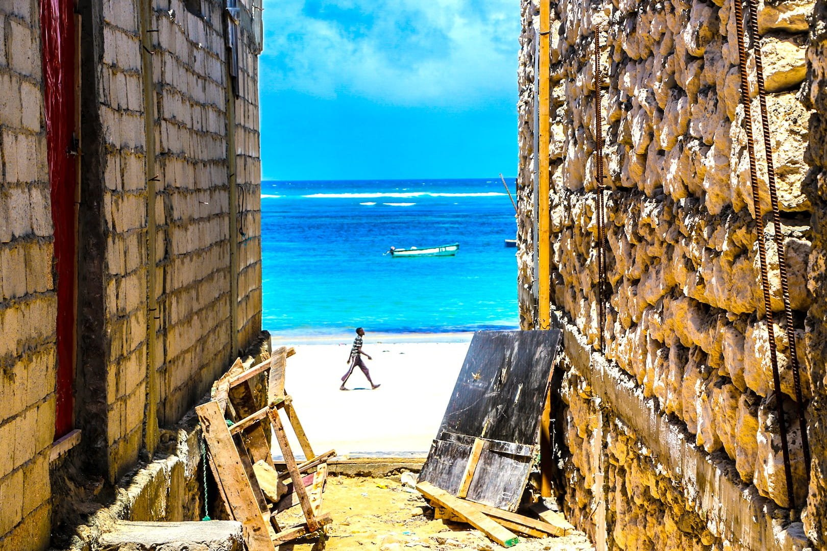 View of the ocean through a construction site between two buildings, with a person walking on the beach and a boat floating on the water in the distance.