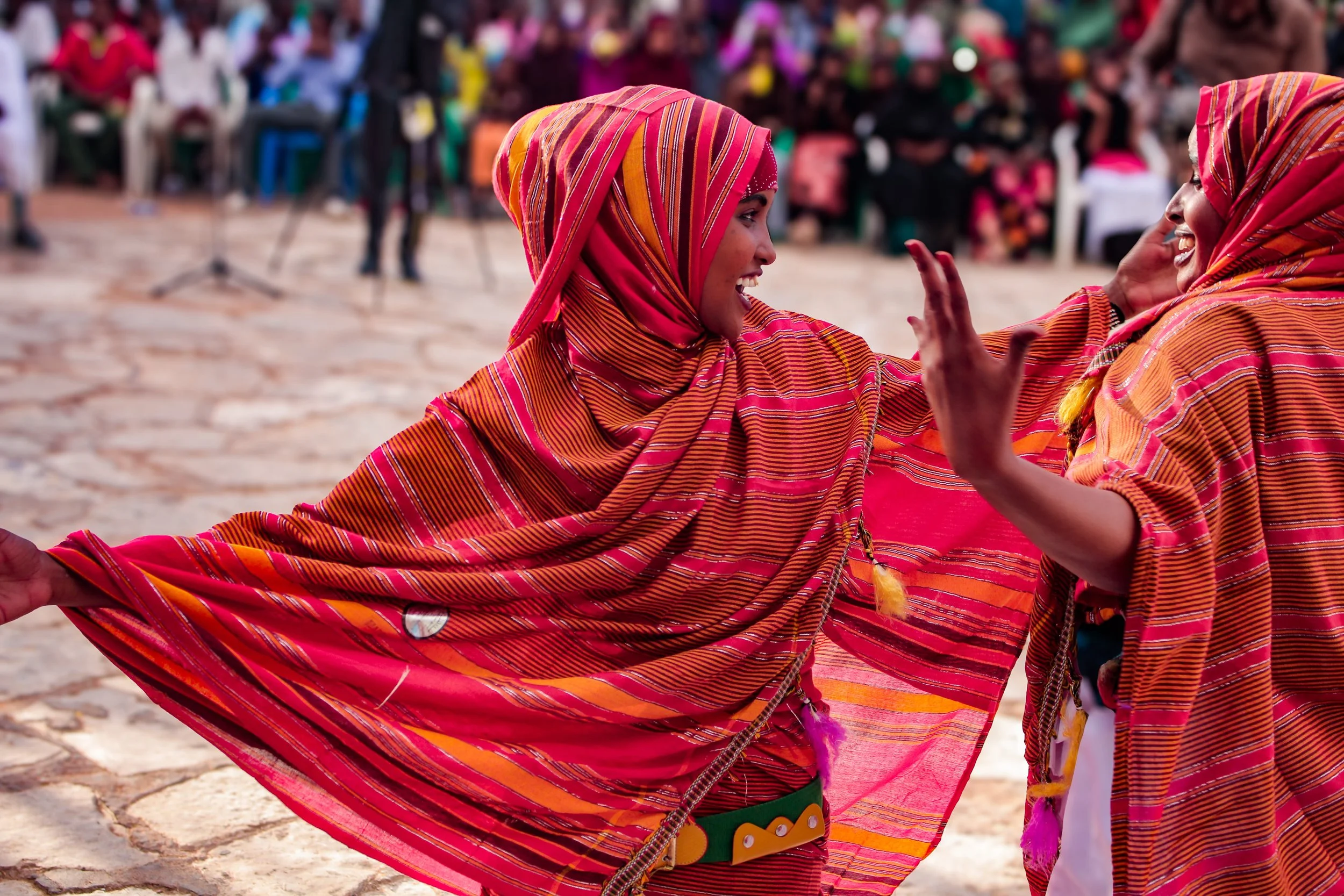 Two women in bright pink and orange traditional clothing dancing and smiling at an outdoor cultural event, with a blurred crowd in the background.