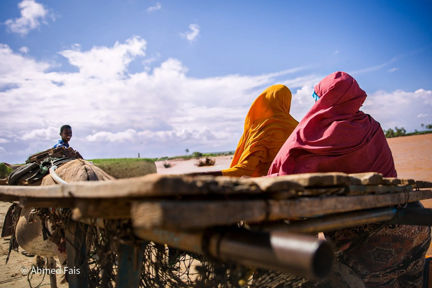 Two women wearing headscarves, one pink and one orange, sitting on a wooden cart. A smiling boy is in the background near a donkey, under a partly cloudy blue sky.
