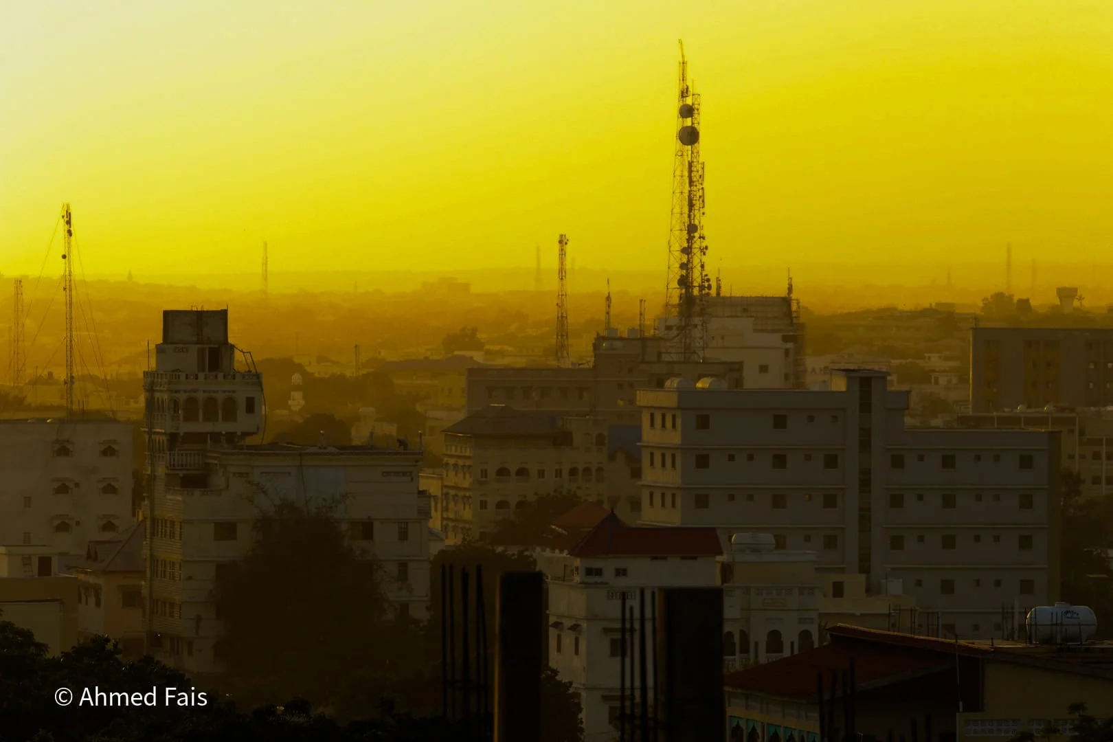 Cityscape during sunset with multiple buildings and communication towers, bathed in yellow light.