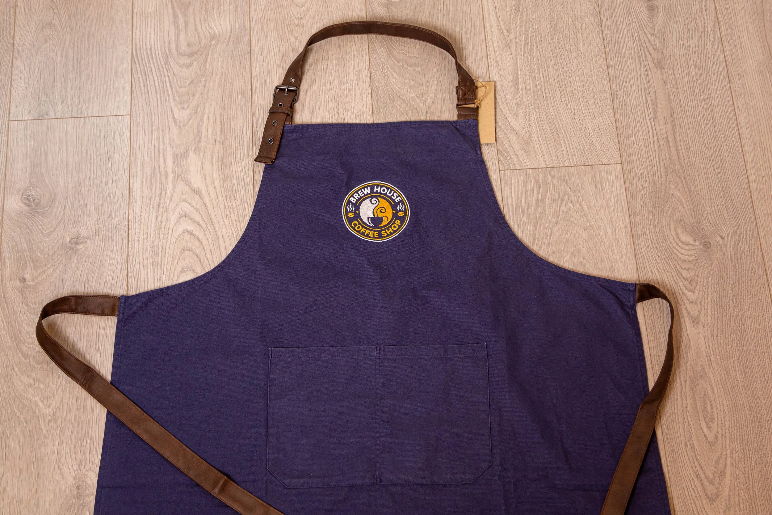 Blue apron with a Brew House Coffee Shop logo, brown leather straps on the neck and waist, laid flat on a wooden floor.