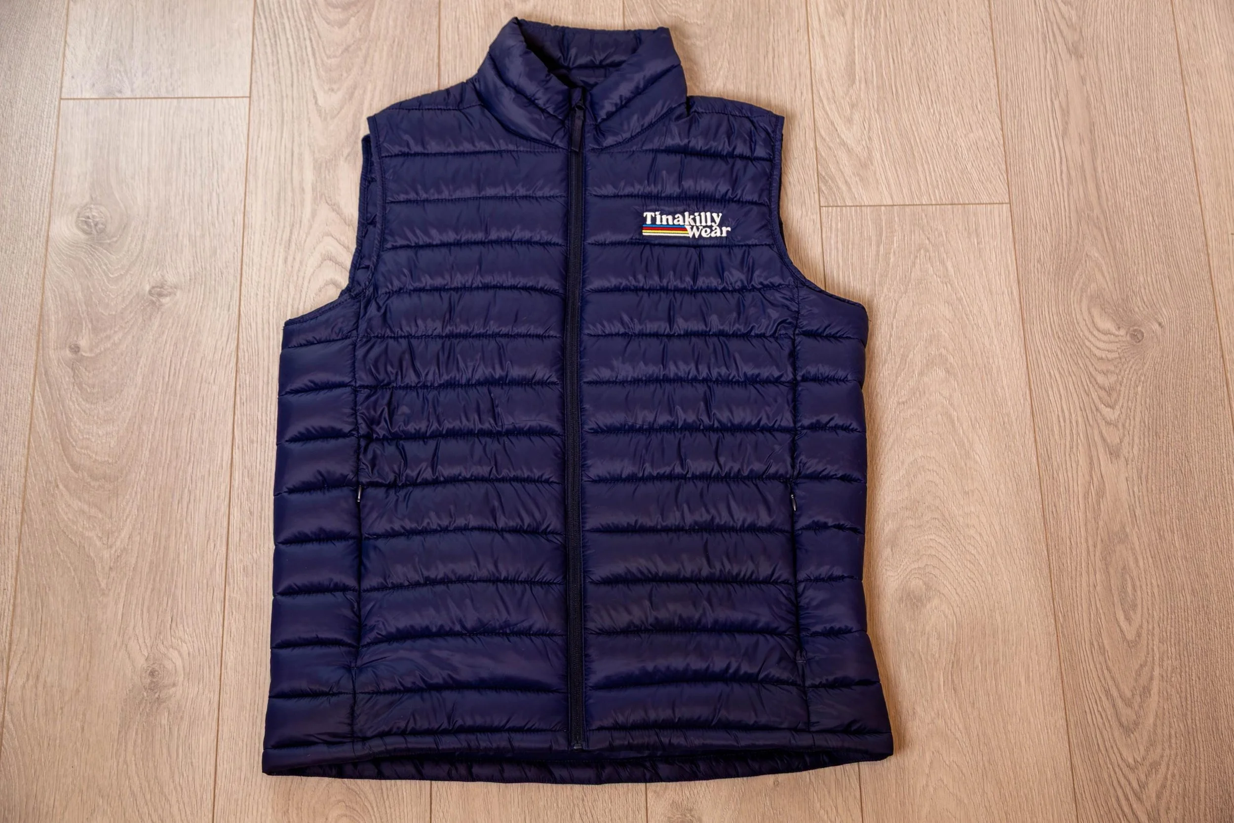 A navy blue quilted vest with a front zipper, placed on a wooden floor. The vest has an embroidered logo that reads 'Tina Kelly Wear'.