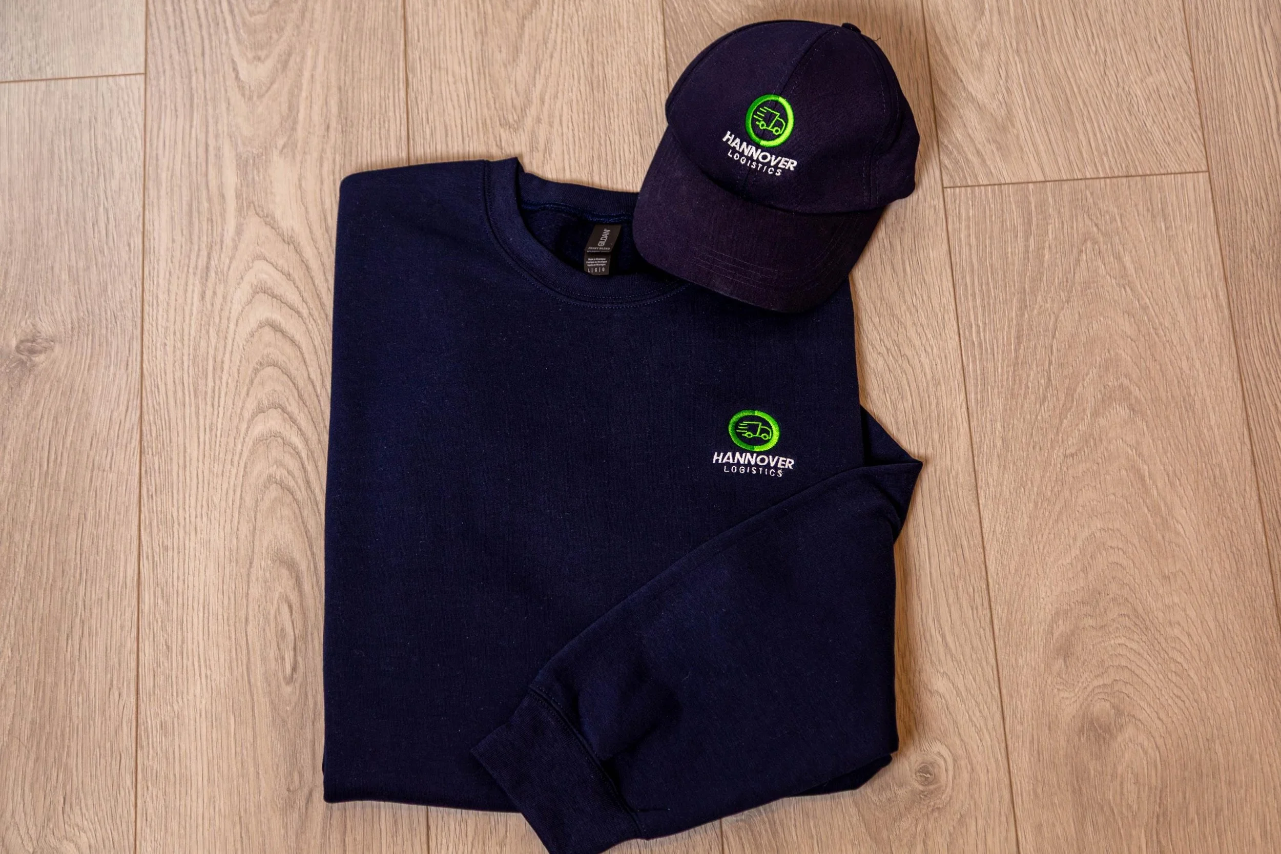 Black sweatshirt and cap with green and white logo of Hannover Logistics on a wooden floor.