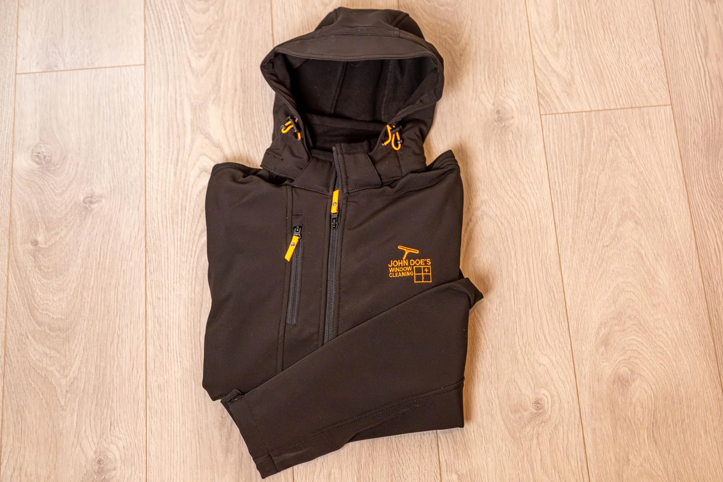 Black hoodie with a logo for John's Window Cleaning, lying on a light-colored wooden floor.