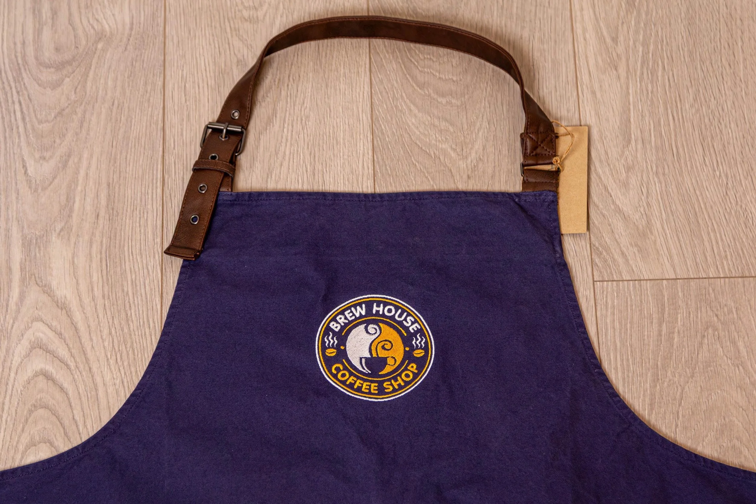 Blue apron with a Brew House Coffee Shop logo and a brown leather strap, lying on a wooden floor.