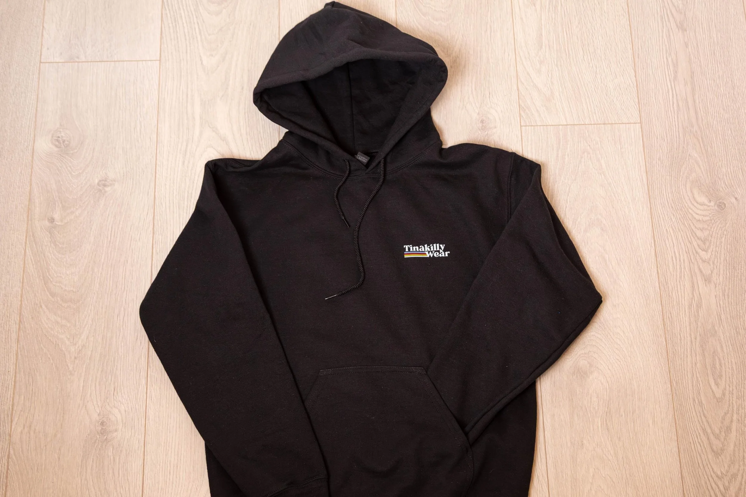 Black hoodie with 'Tinakilly Wear' logo on the chest, laid flat on a wood floor.