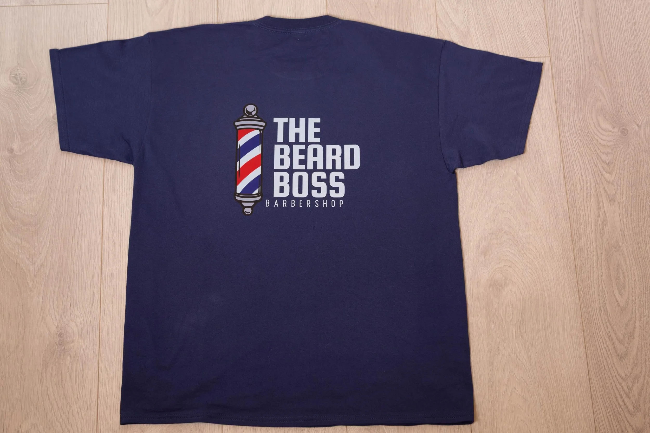 Navy blue T-shirt with a barber pole graphic and the words 'The Beard Boss Barbershop' printed on the front, laid flat on a wooden surface.