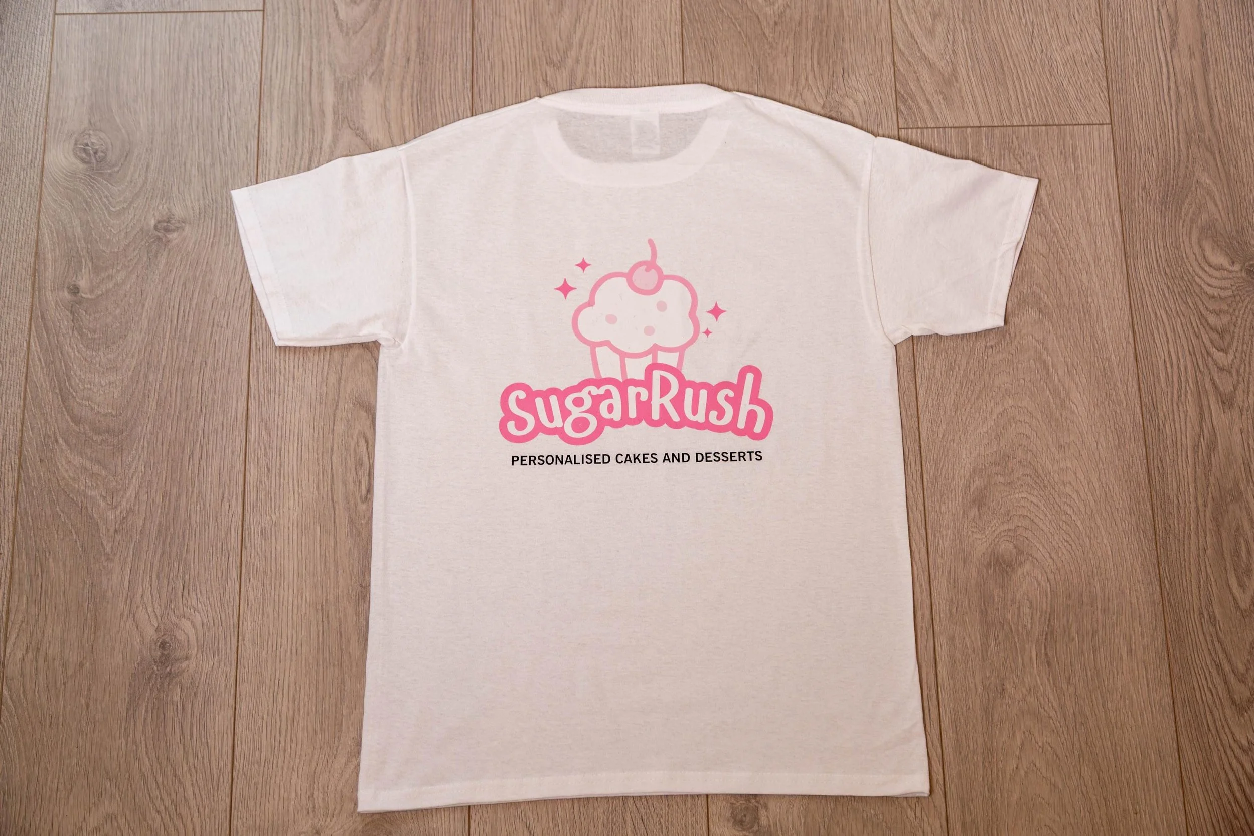 A white T-shirt with a pink logo of a cupcake and the words 'Sugar Rush' on it, along with the tagline 'Personalised Cakes and Desserts'.