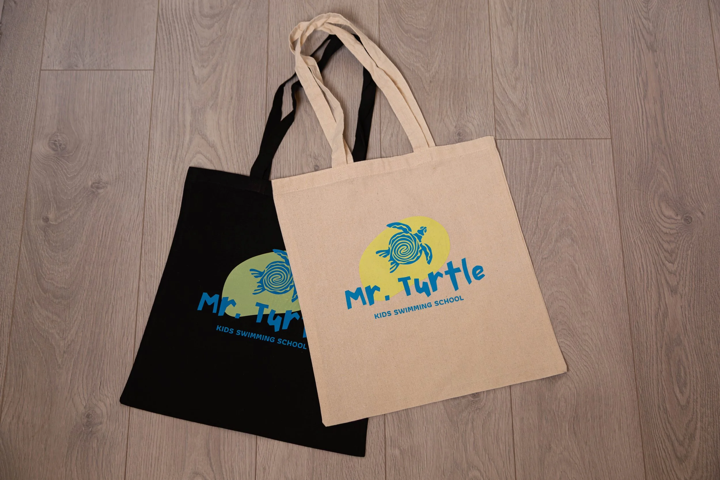 Two tote bags, one black and one beige, with a blue and yellow logo of a turtle and the text "Mr. Turtle Kids Swimming School," placed on a wooden floor.