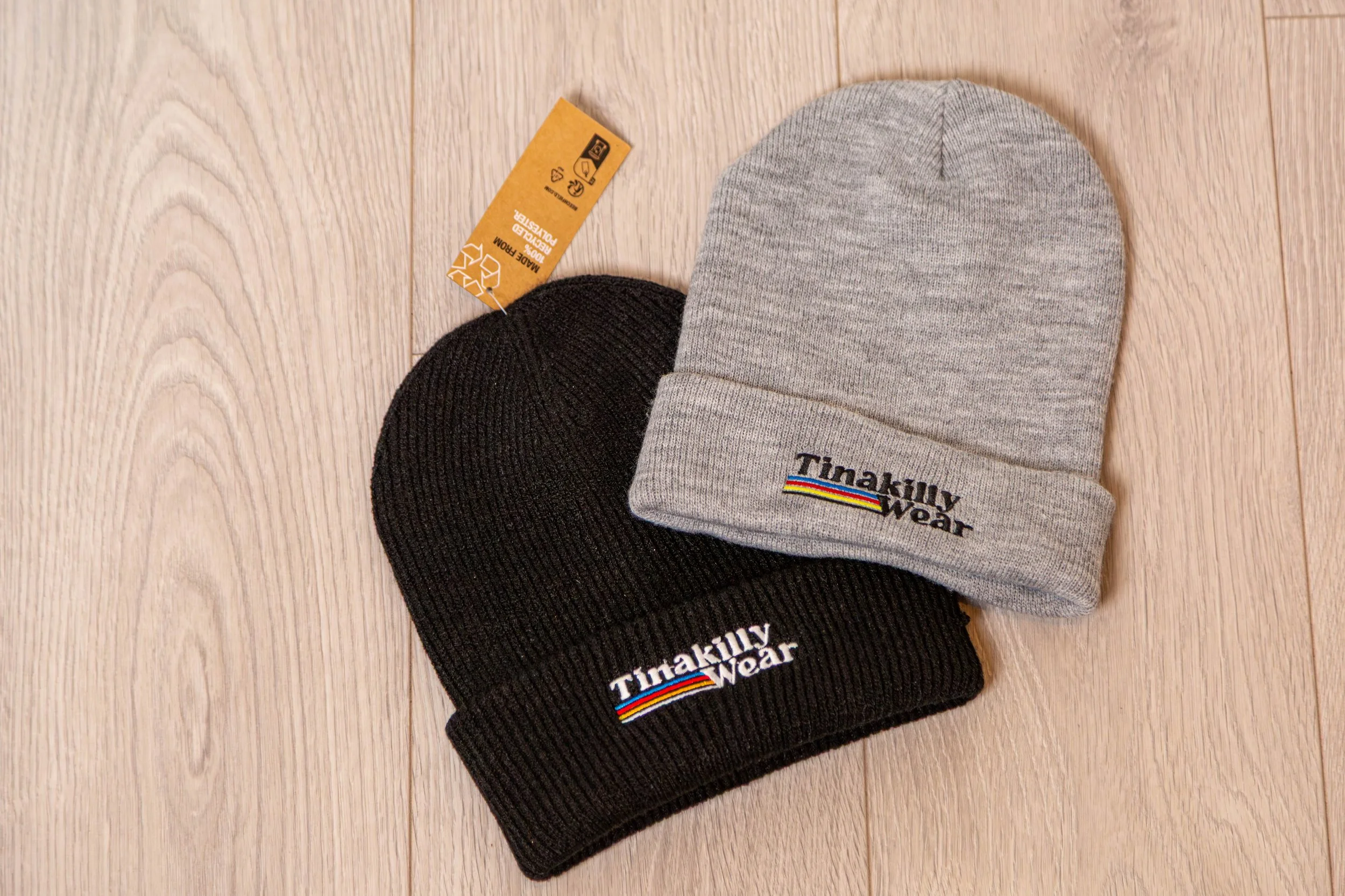 Two beanies, one black and one gray, with 'Tinalilly Wear' embroidered logos, placed on a wooden floor.