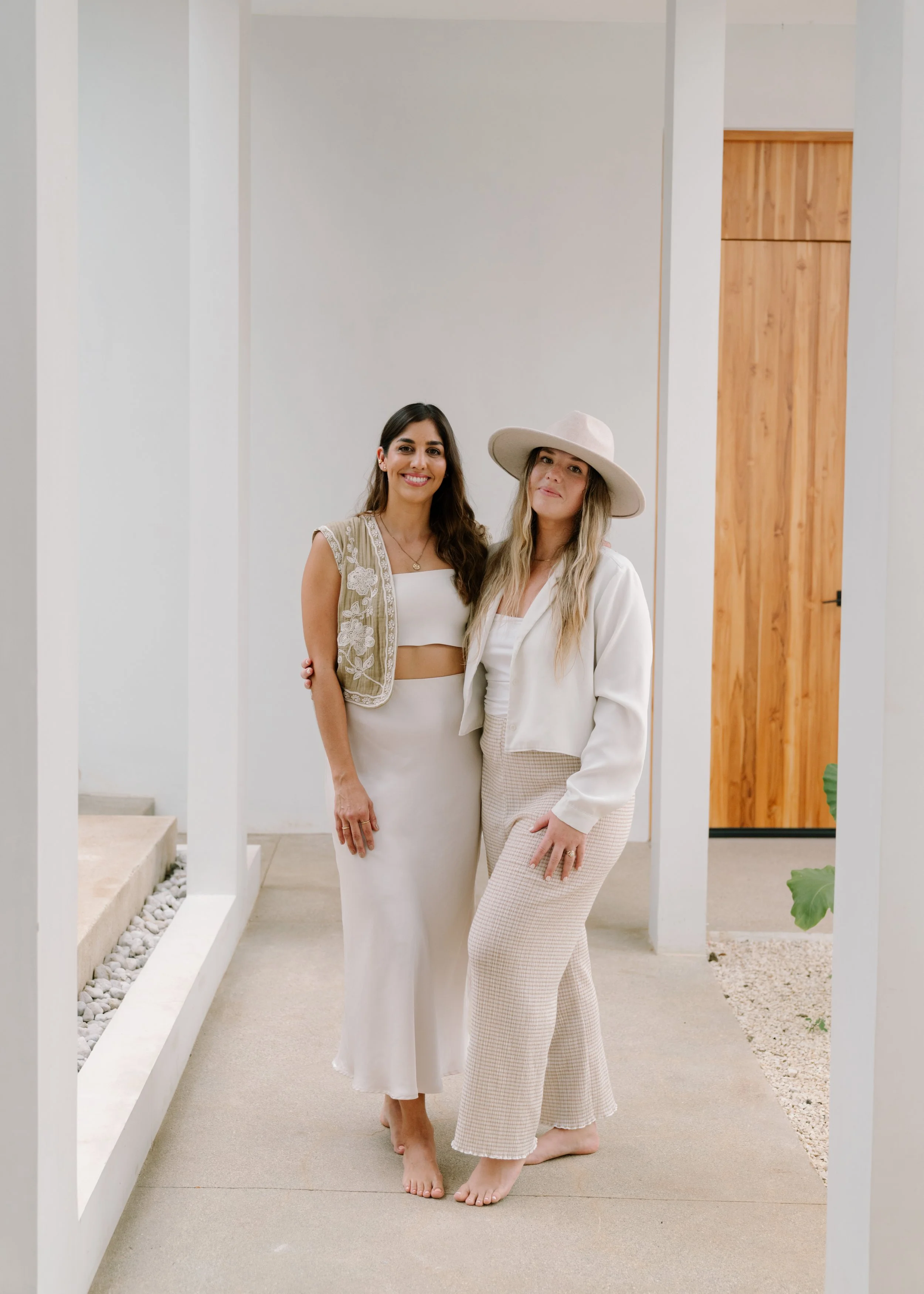 Two women standing together indoors, smiling, with one wearing a white dress and the other in a white jacket, beige patterned pants, and a wide-brimmed hat.