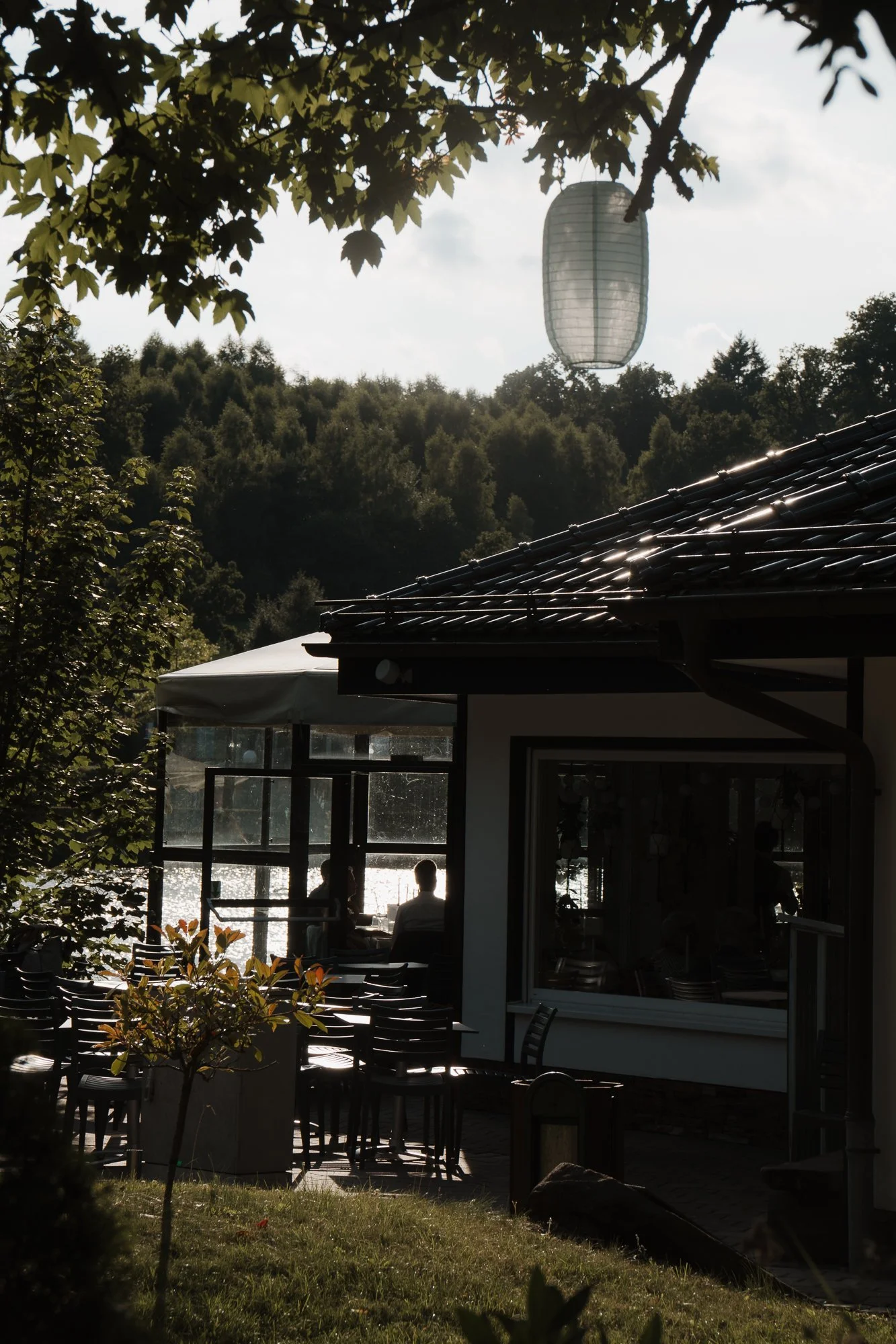 Schattenhafte Szene eines Restaurants am Wasser mit Blick auf einen bewaldeten Hügel im Hintergrund, im Vordergrund sind Stühle, Tische und Fenster sichtbar, mit Menschen, die draußen sitzen, während eine Laterne in der Luft schwebt.