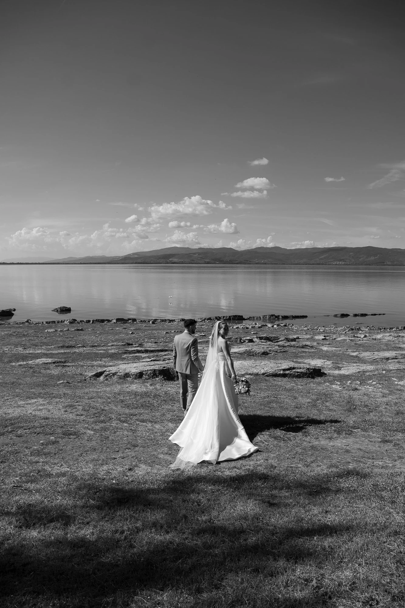 Ein Brautpaar, das am Strand bei Sonnenlicht steht, die Braut trägt ein langes, weißes Brautkleid mit Schleier, der Bräutigam trägt einen Anzug, im Hintergrund ein ruhiger See und Berge, in Schwarz-Weiß