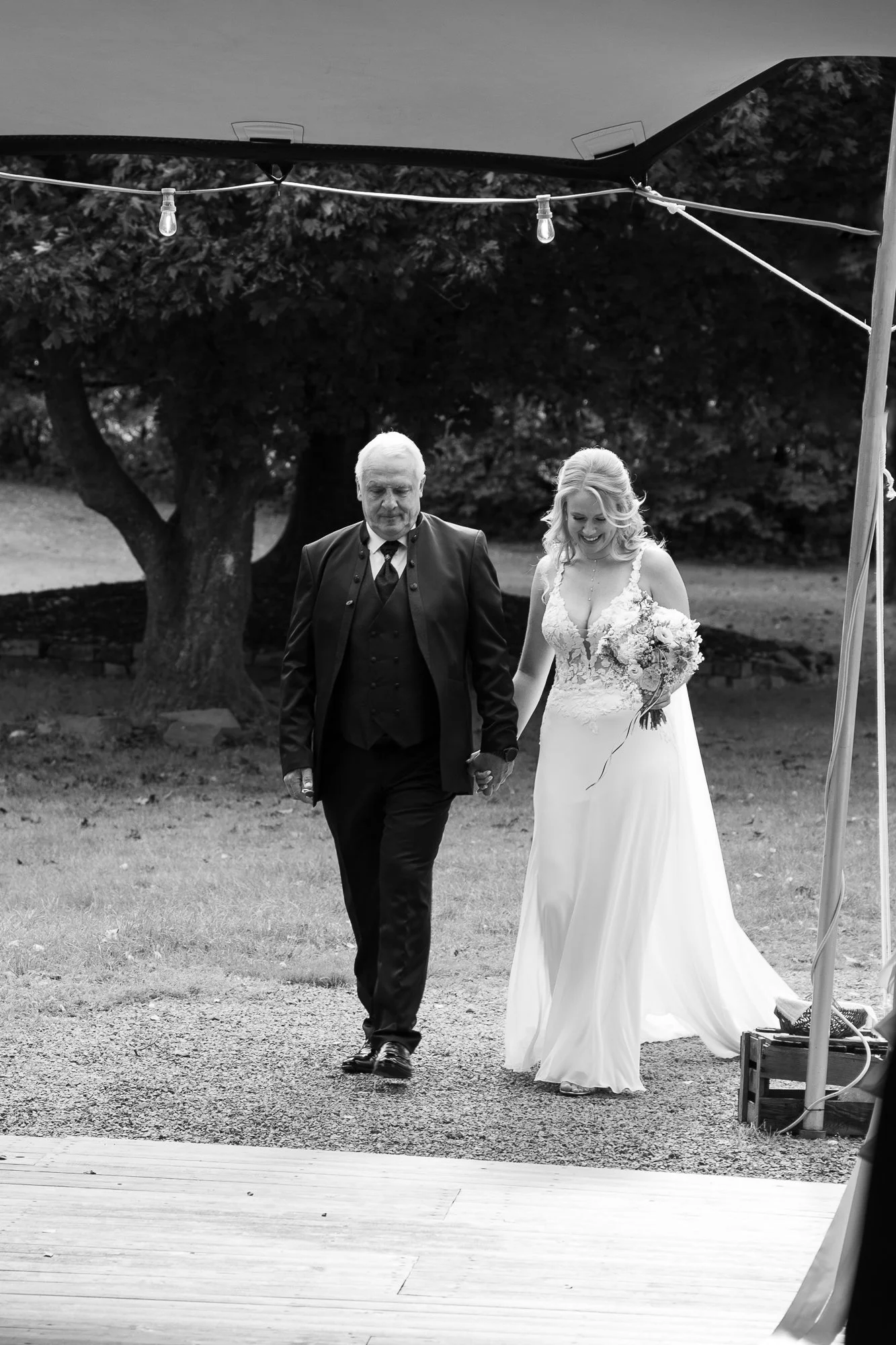 Braut in weißem Kleid mit Blumenstrauß, geht Hand in Hand mit älterem Mann im Anzug bei einer Hochzeit im Freien.
