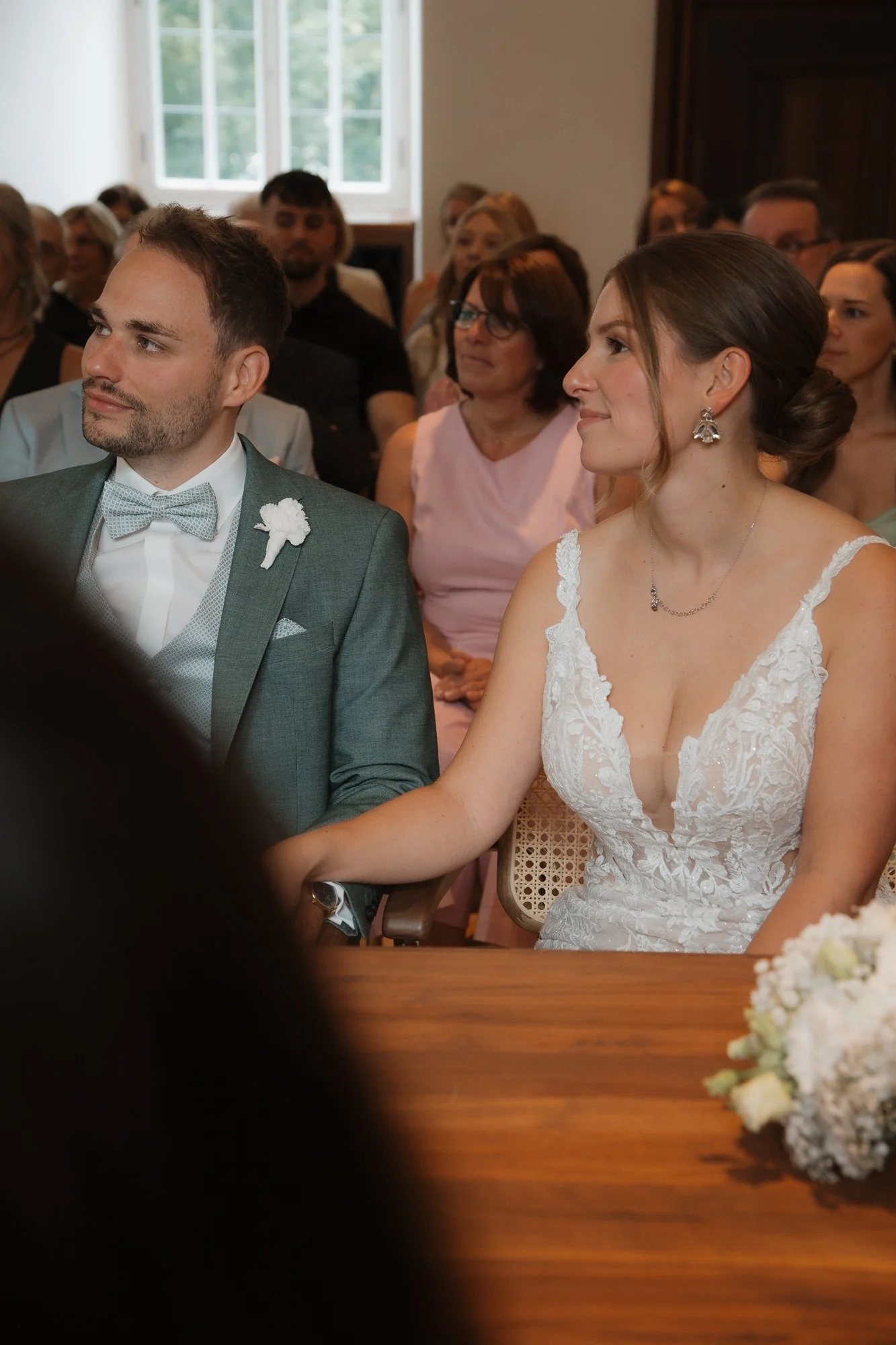 Ein Brautpaar bei einer standesamtlichen Hochzeit, sitzend an einem Tisch, umgeben von Gästen in einem festlich dekorierten Raum.