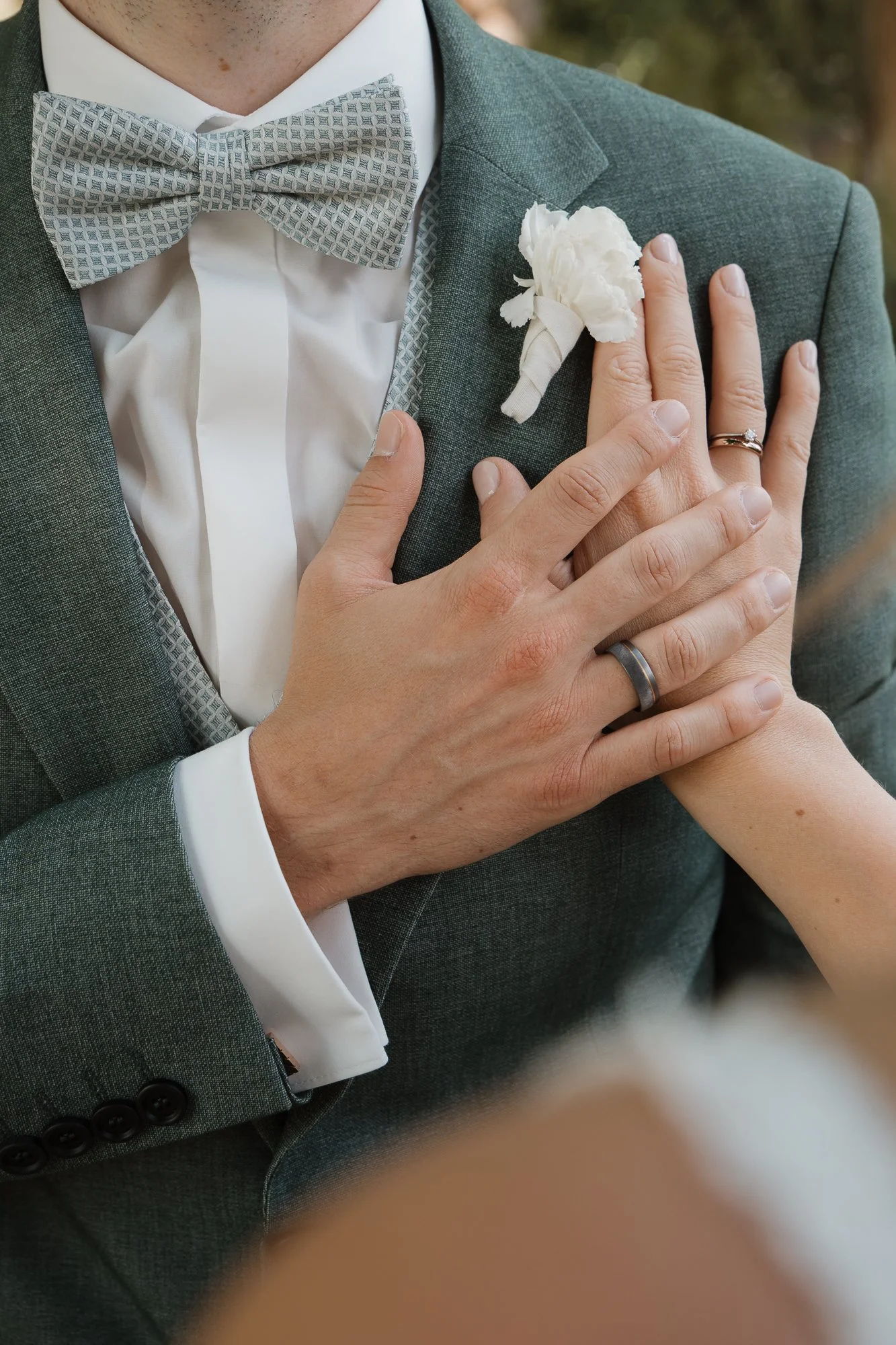 Nahaufnahme eines Bräutigams in einem grauen Anzug mit weißem Hemd und kariertem Fliege, der eine weiße Blumenkrawatte trägt. Die Hand einer Braut mit Ehering berührt die Brusttasche des Anzugs.