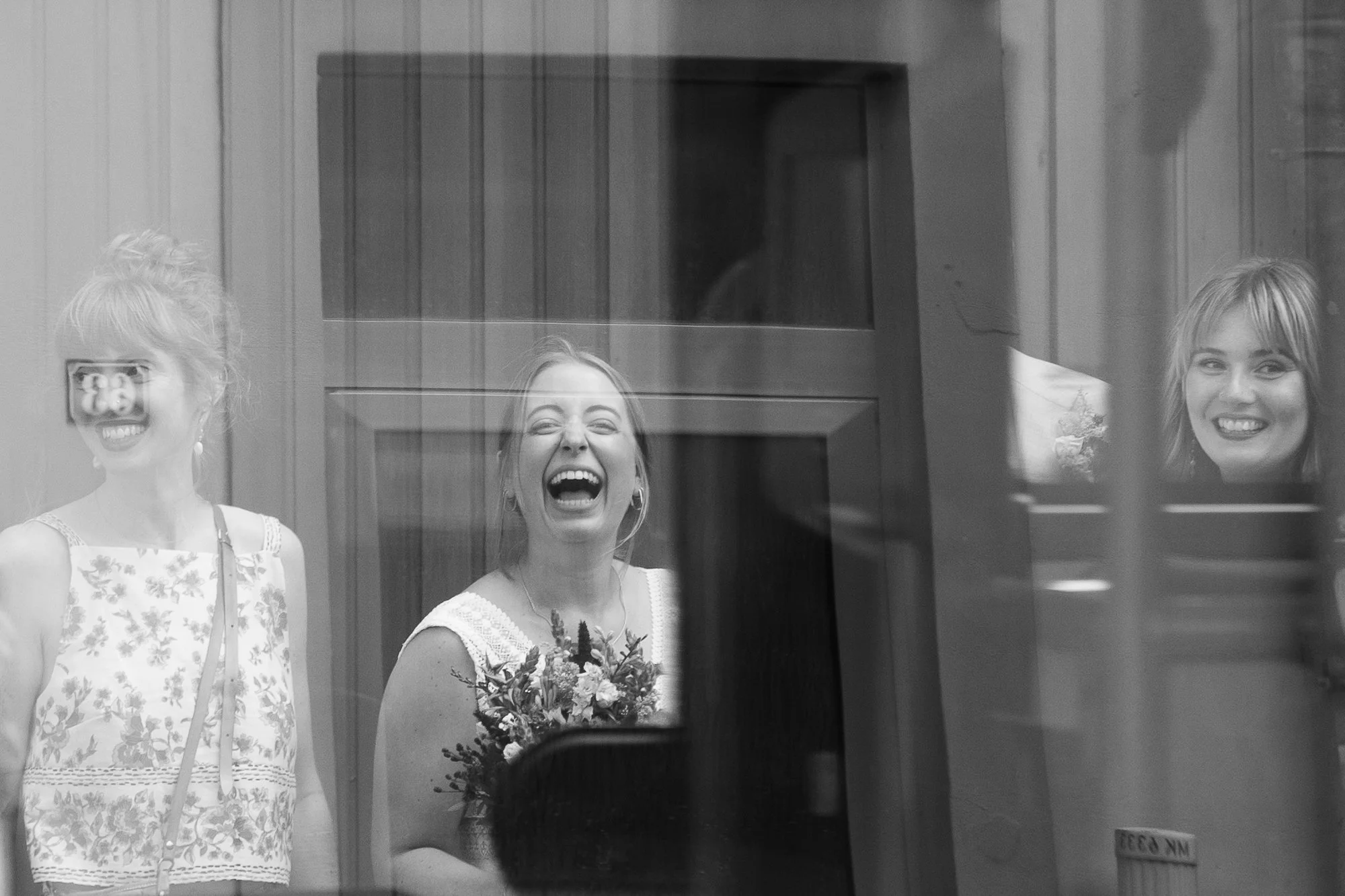 Drei lachende Frauen, die durch eine Fensterscheibe gesehen werden, während eine von ihnen ein Blumenstrauß hält.