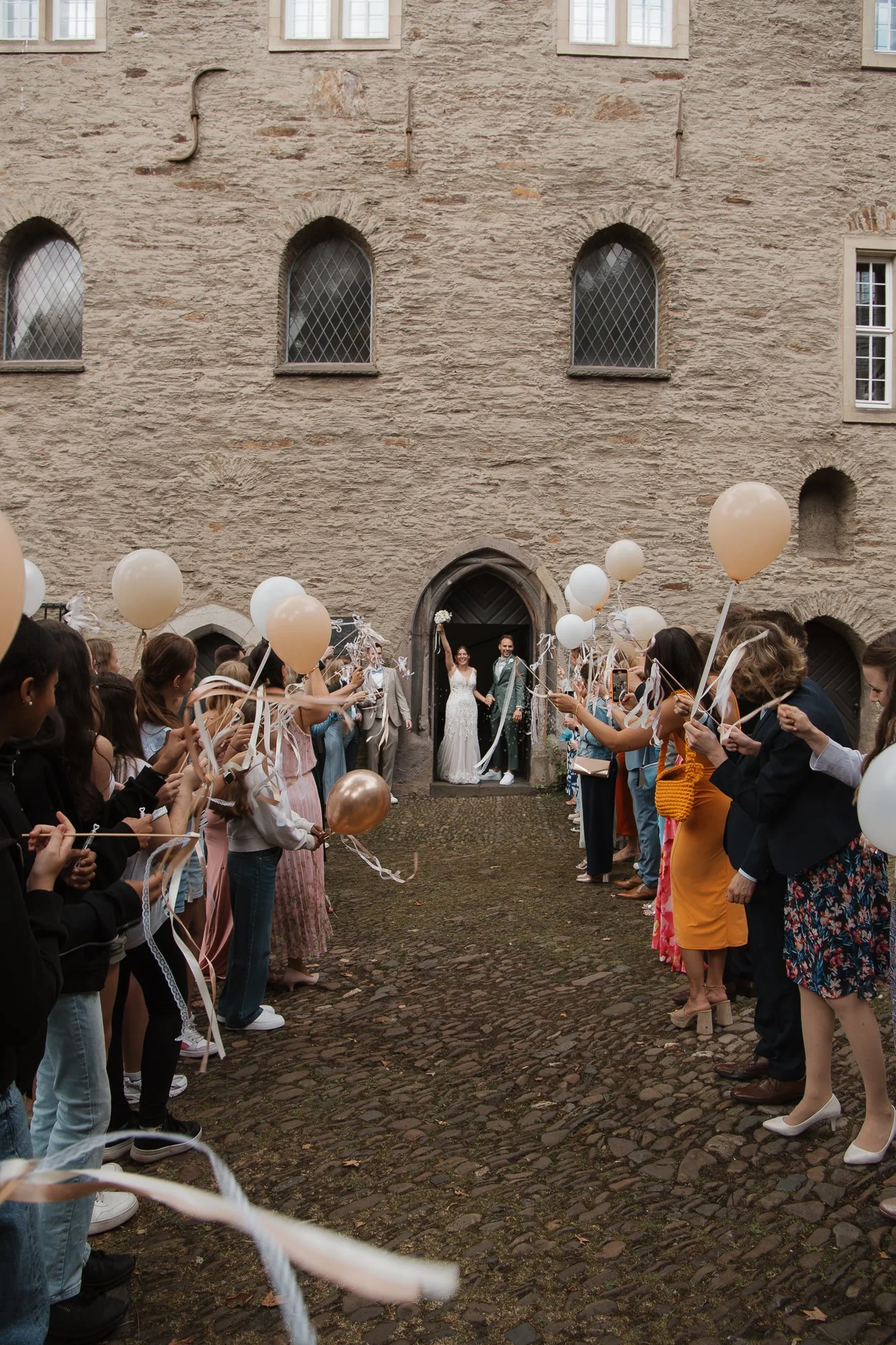 Hochzeitspaar steht vor einem alten Steinmauer, umgeben von Gästen mit Luftballons, die ihnen gratulieren.