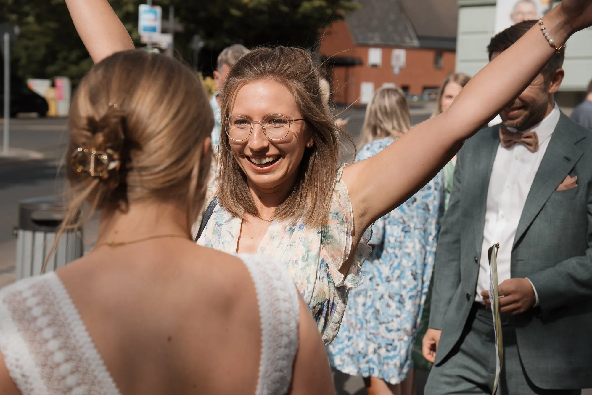 Eine Frau mit Brille freut sich, im Gespräch mit einer anderen Frau, während ein Mann im Anzug im Hintergrund steht.