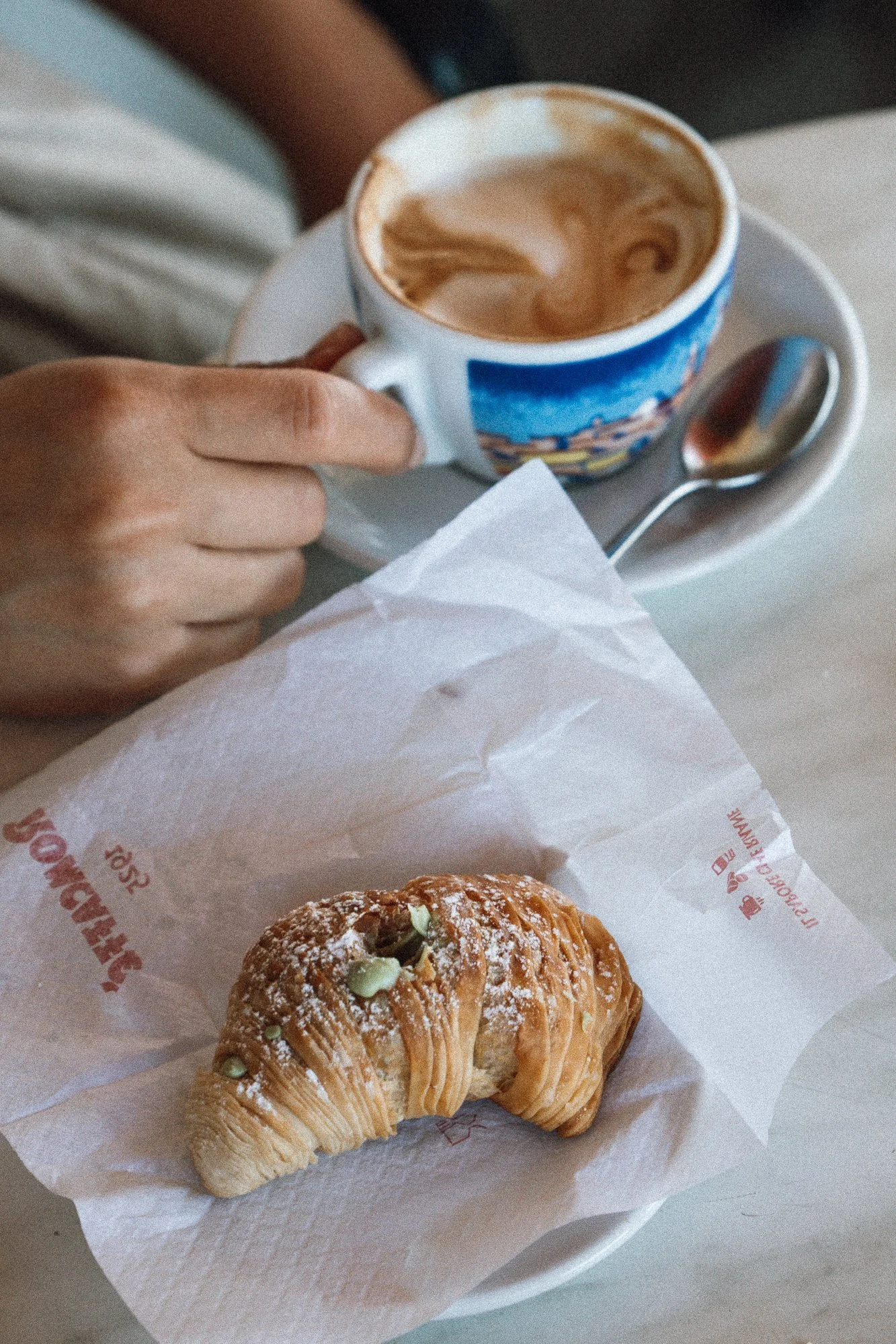 Eine Tasse Cappuccino mit Latte Art auf einem weißen Teller, daneben ein karamellisiertes Croissant auf Pergamentpapier.