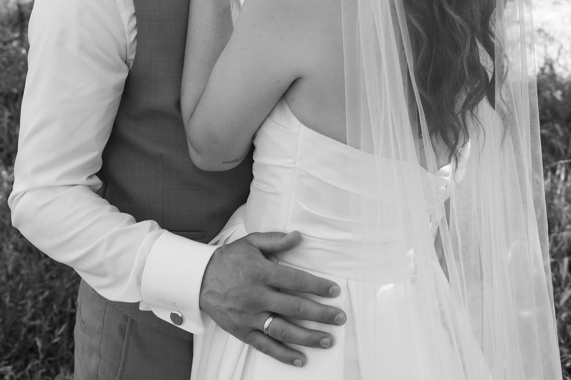 Nahaufnahme eines Brautpaares, wobei der Bräutigam eine Hand auf die Taille der Braut legt. Das Hochzeitskleid der Braut ist sichtbar, ebenso der Schleier. Der Hintergrund ist unscharf. Das Bild ist in Schwarz-Weiß.