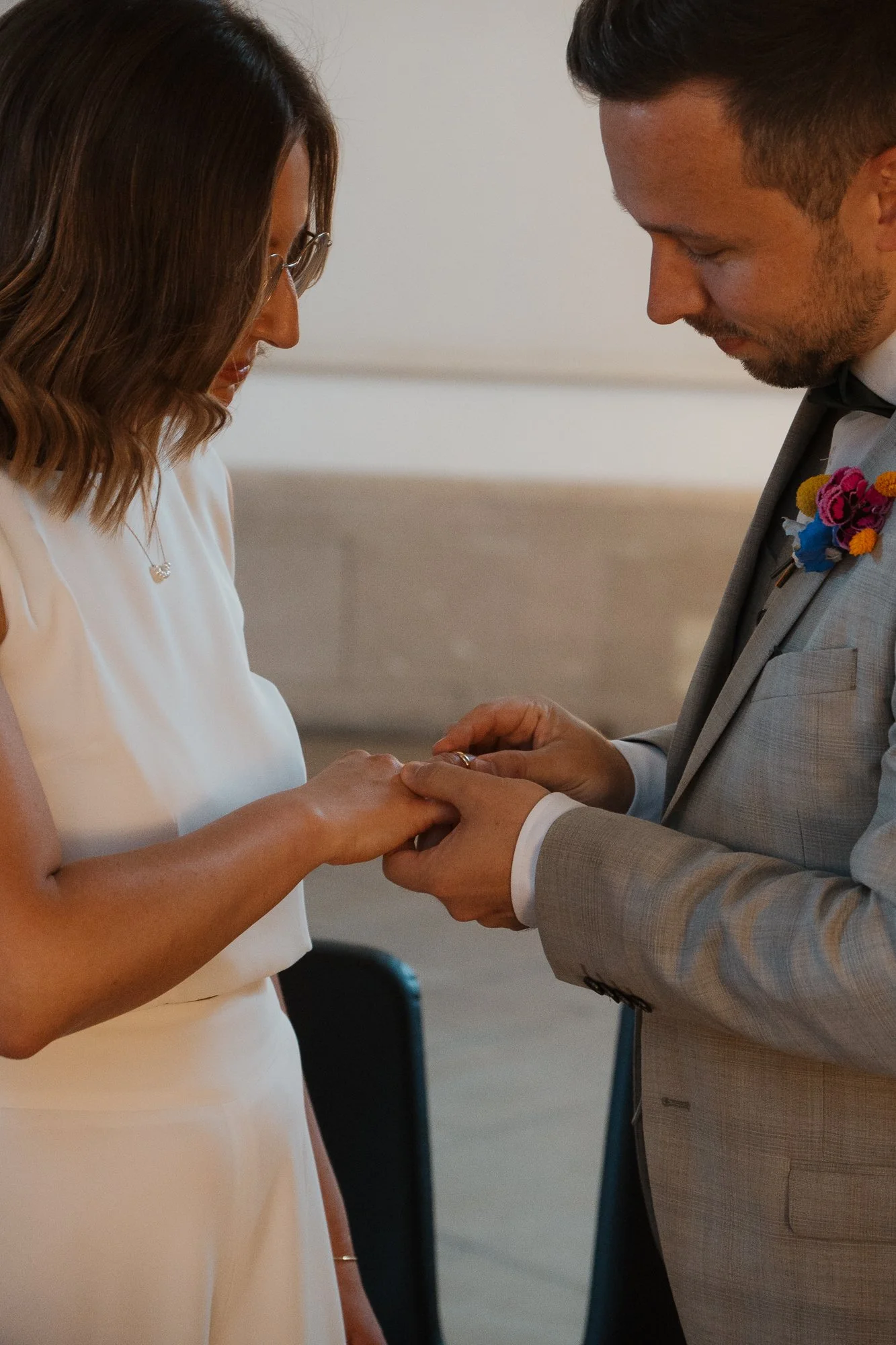 Ein Mann und eine Frau bei einer Eheschließung, der Mann steckt der Frau einen Ring an den Finger, die Frau trägt ein weißes Kleid, der Mann trägt einen Anzug mit einer Blumenbrosche.
