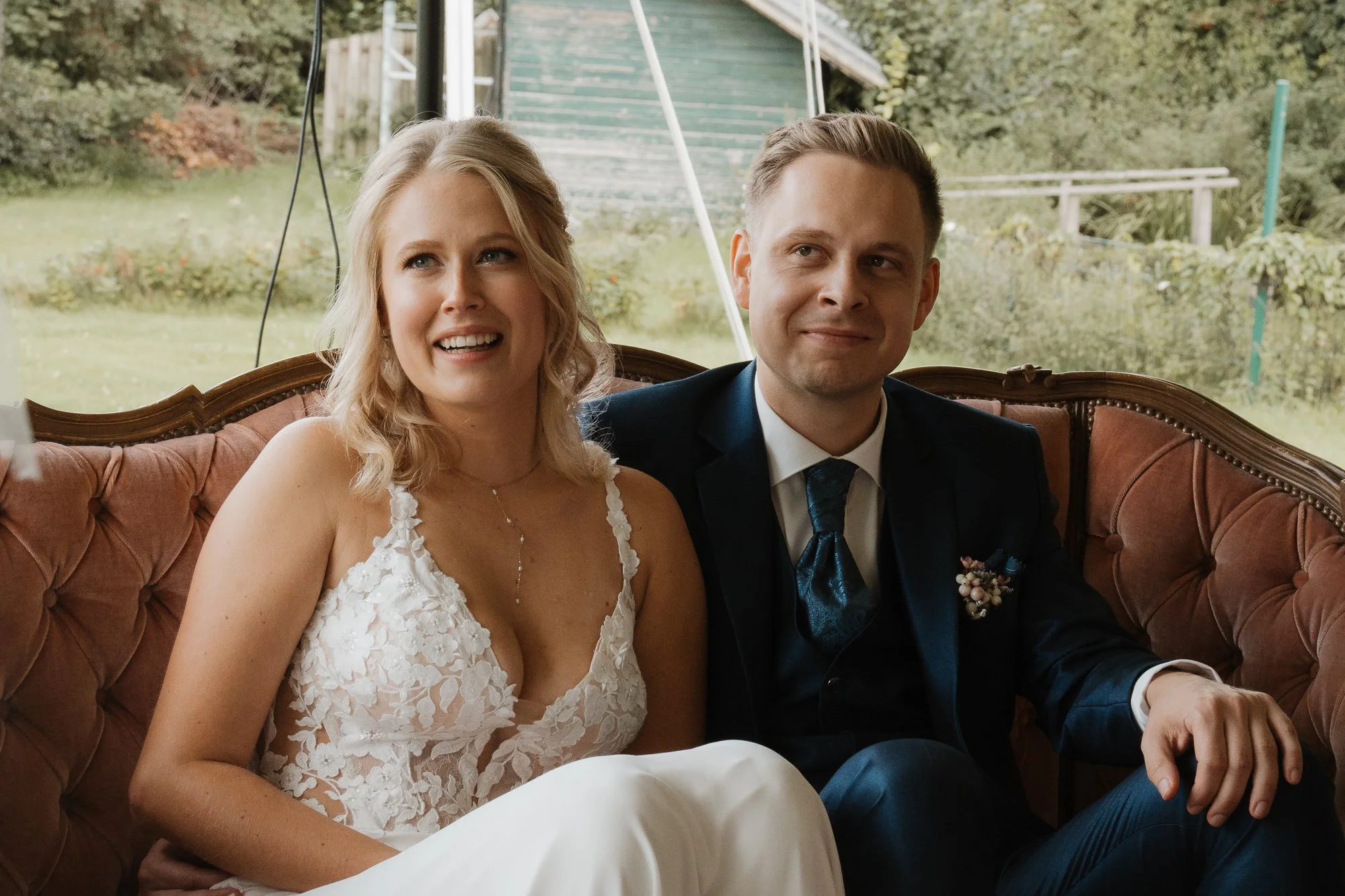 Ein lachendes Brautpaar sitzt auf einem alten Sofas in einem Garten an einem Hochzeitstag.