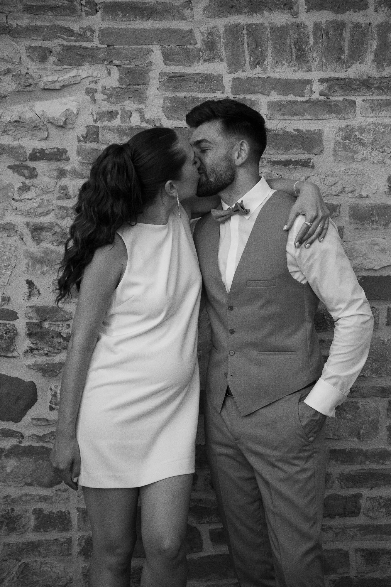 Ein Mann und eine Frau küssen sich vor einer Ziegelmauer, sie trägt ein weißes Kleid, er einen Anzug mit Weste und Fliege.