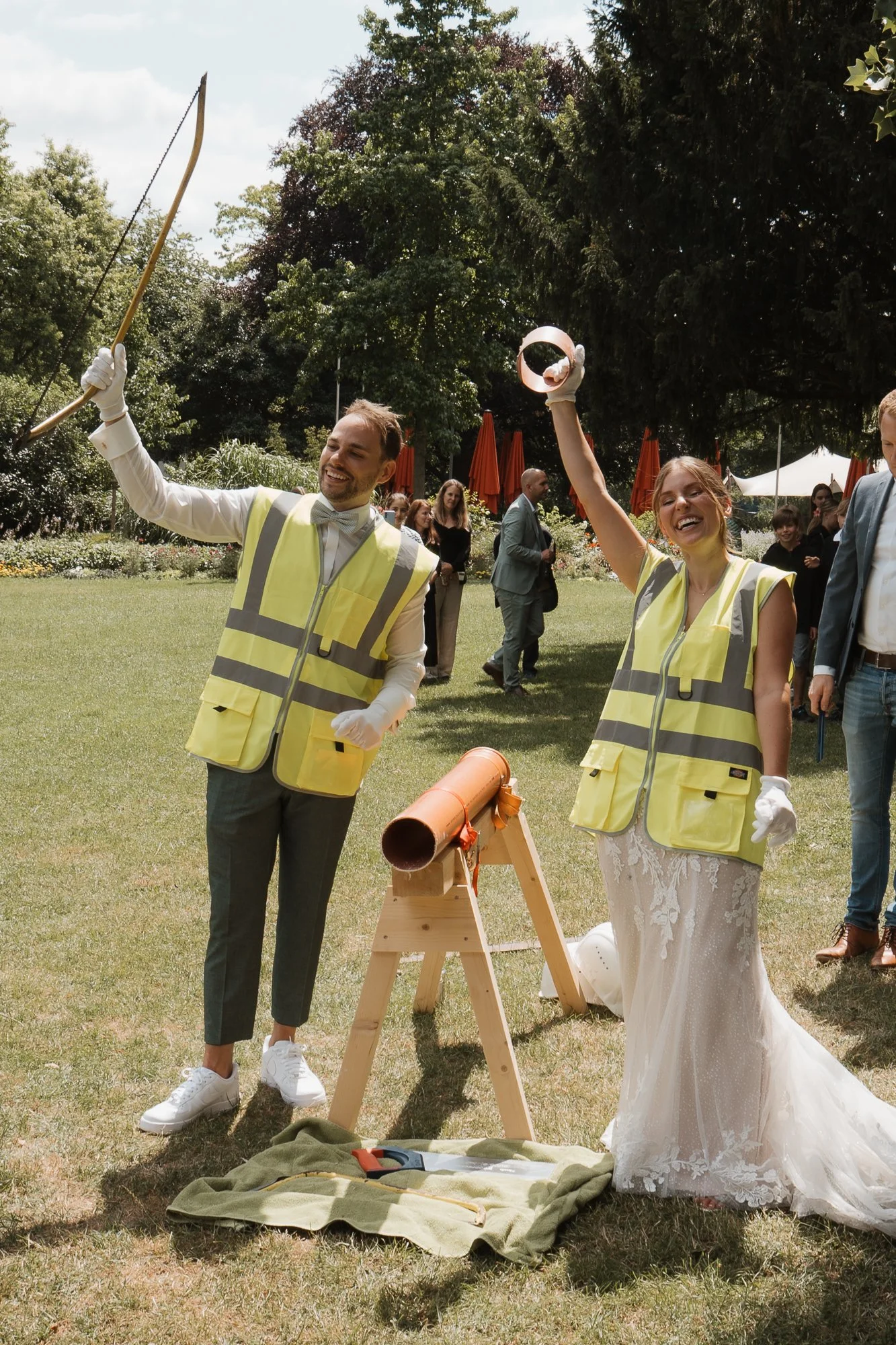 Gruppe von Hochzeitsgästen in Warnwesten, die am Hochzeitstag mit einem Teleskop auf dem Rasen im Park eine Rakete ins All schießen, lächelnd und spielerisch, bei sonnigem Wetter, im Hintergrund weitere Gäste und Bäume.