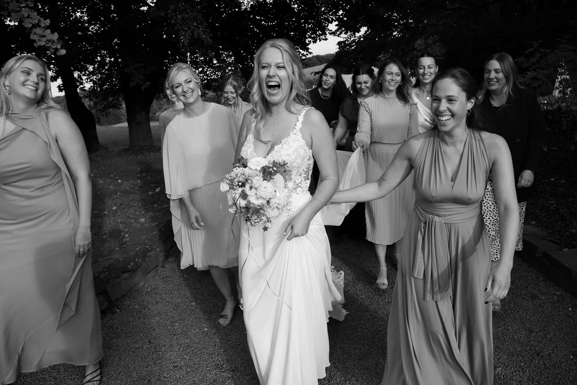 Schwarz-Weiß-Foto einer Hochzeitsgesellschaft, die eine Braut in einem Brautkleid mit Blumenstrauß zeigt, umgeben von lachenden Frauen in Sommerkleidern im Freien unter Bäumen.