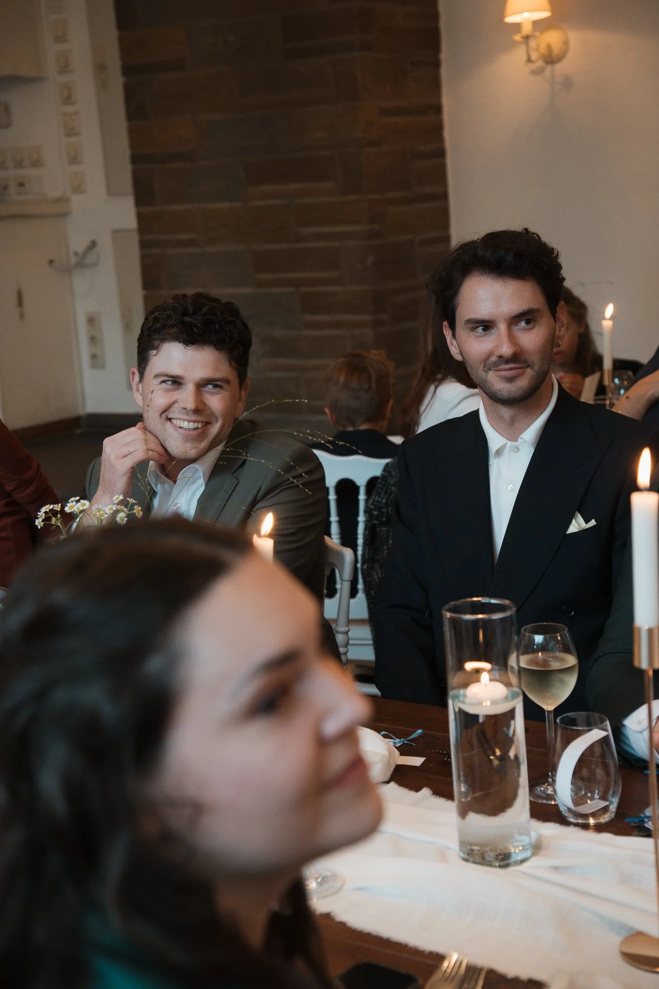 Zwei Männer sitzen an einem festlich gedeckten Tisch bei einer Veranstaltung, lachen und schauen in unterschiedliche Richtungen, begleitet von Kerzenlicht und einem Glas Weißwein. Im Hintergrund weitere Gäste und eine Wand mit Holzverkleidung.