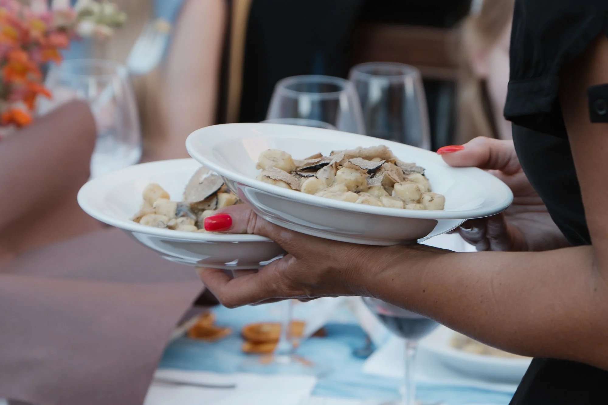 Nahaufnahme einer Person, die zwei weiße Teller mit einem Gericht aus Trüffeln, Pilzen und Pasta hält. Im Hintergrund sind leere Weingläser und ein gedeckter Tisch sichtbar.